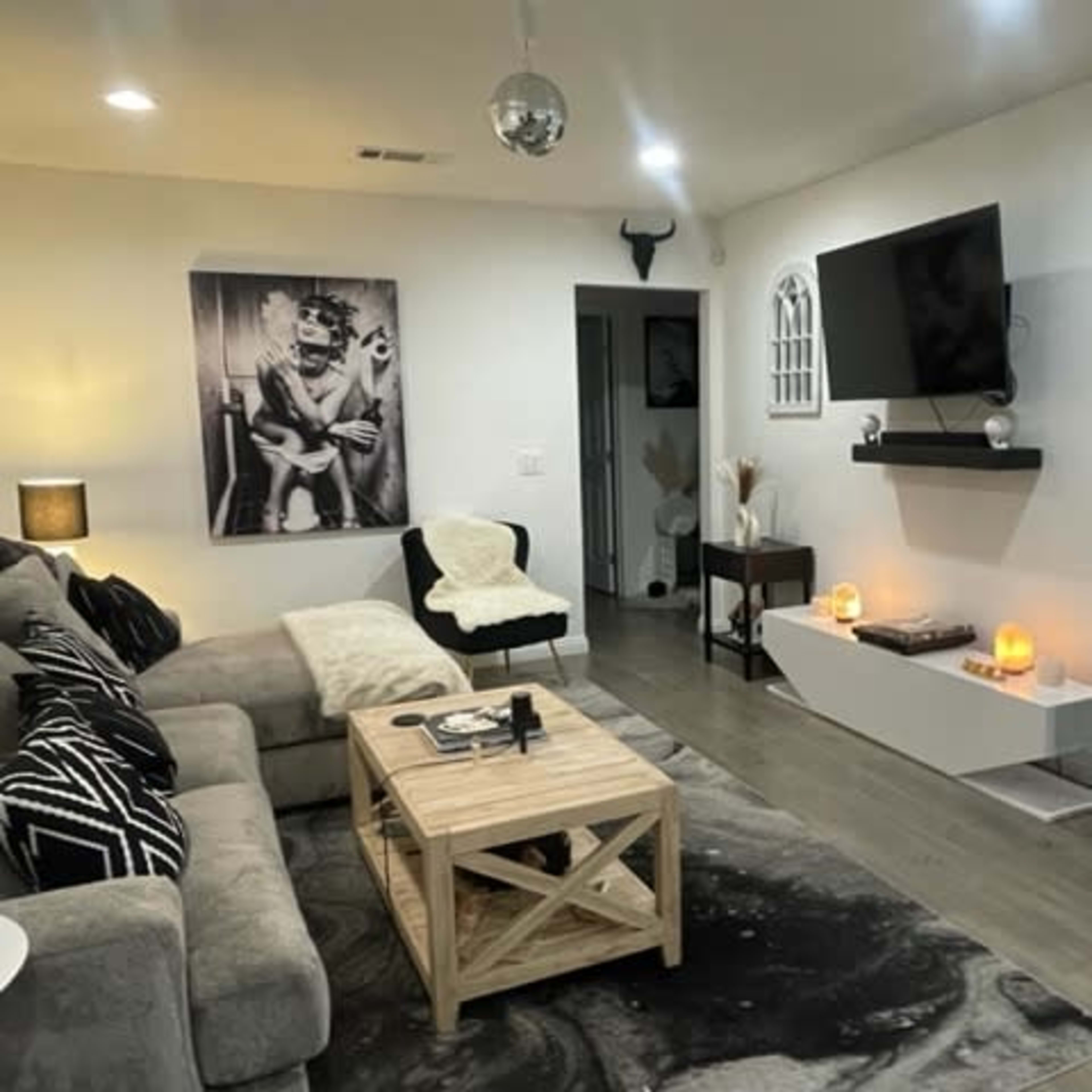 A living room with a gray sectional sofa, a wooden coffee table, a large wall-mounted TV, and decorative accents like candles and framed art.