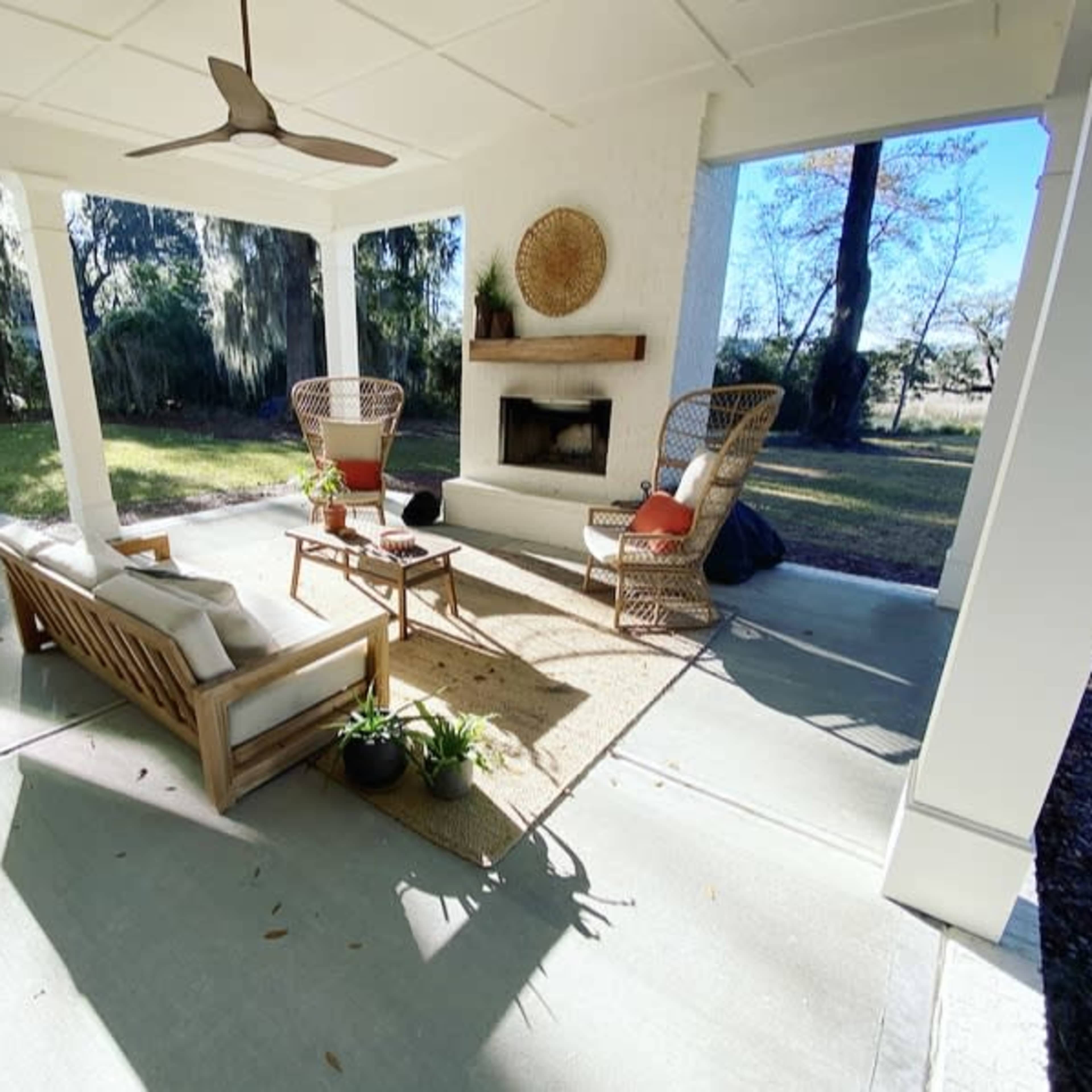 A covered outdoor space featuring a seating arrangement with two chairs, a sofa, a coffee table, and a fireplace, surrounded by greenery.