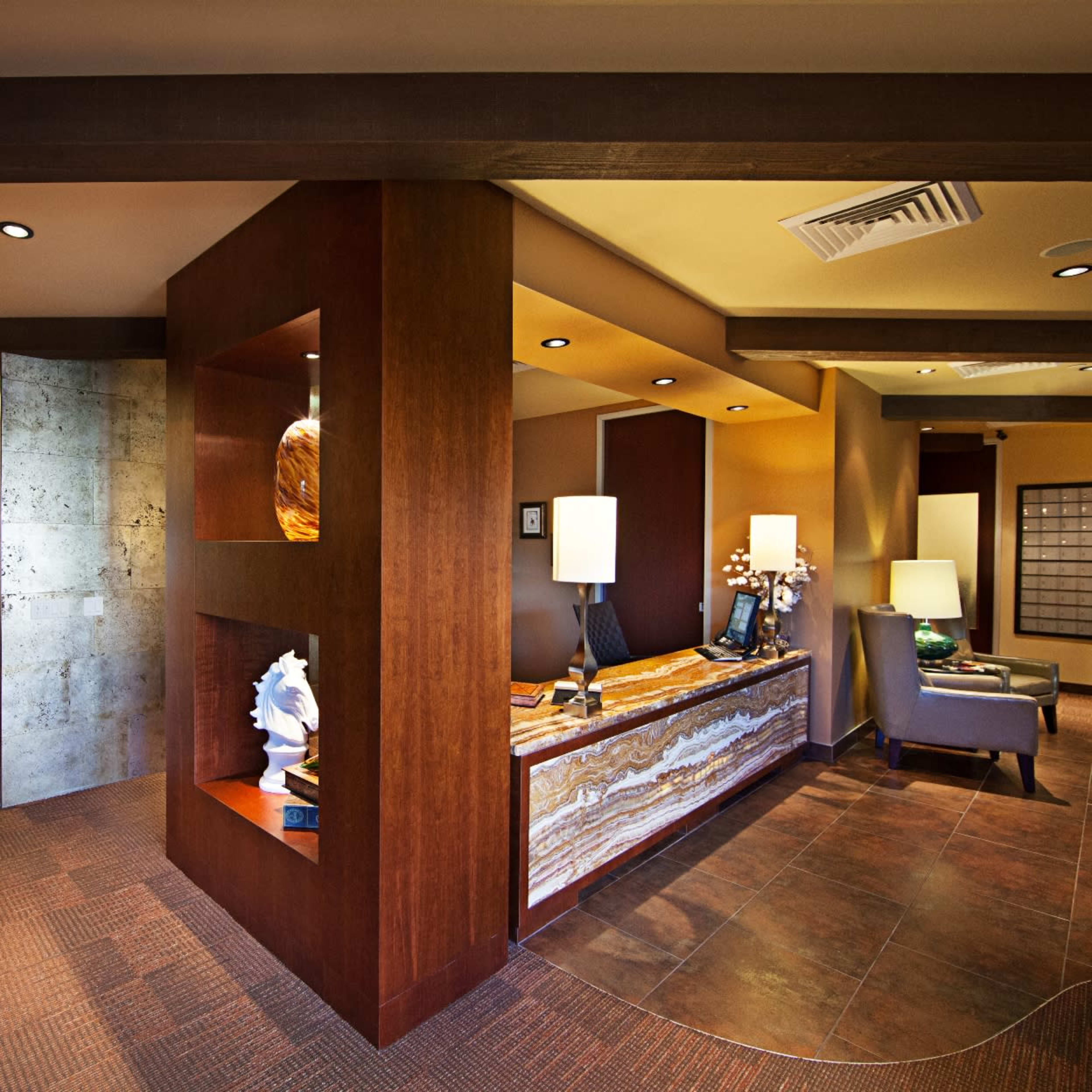 A modern office reception area featuring a wooden reception desk with lamps and a textured wall, with a corridor leading to additional office spaces.