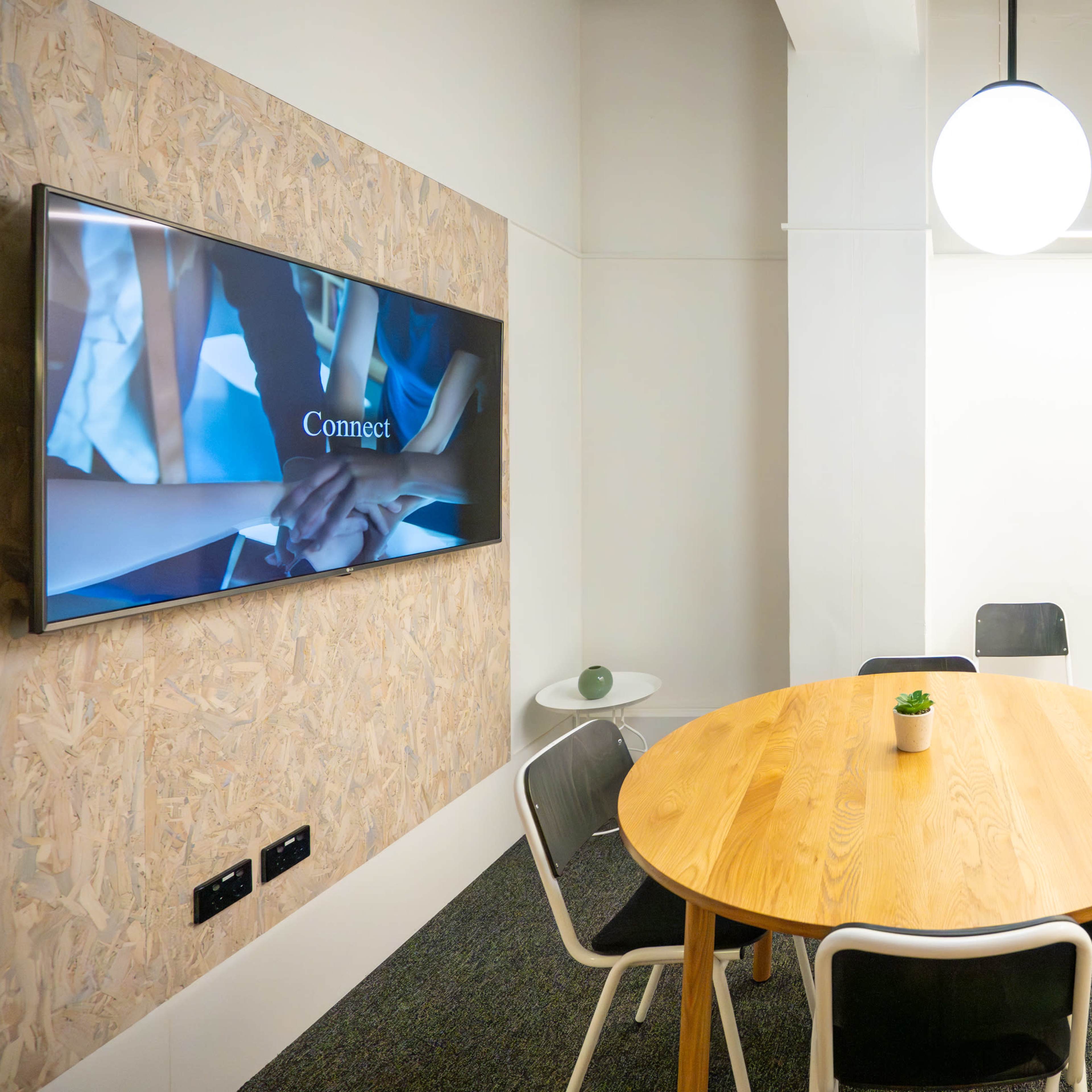 A small conference room features a round wooden table surrounded by black and white chairs, a wall-mounted screen displaying a presentation, and a whiteboard on one side.