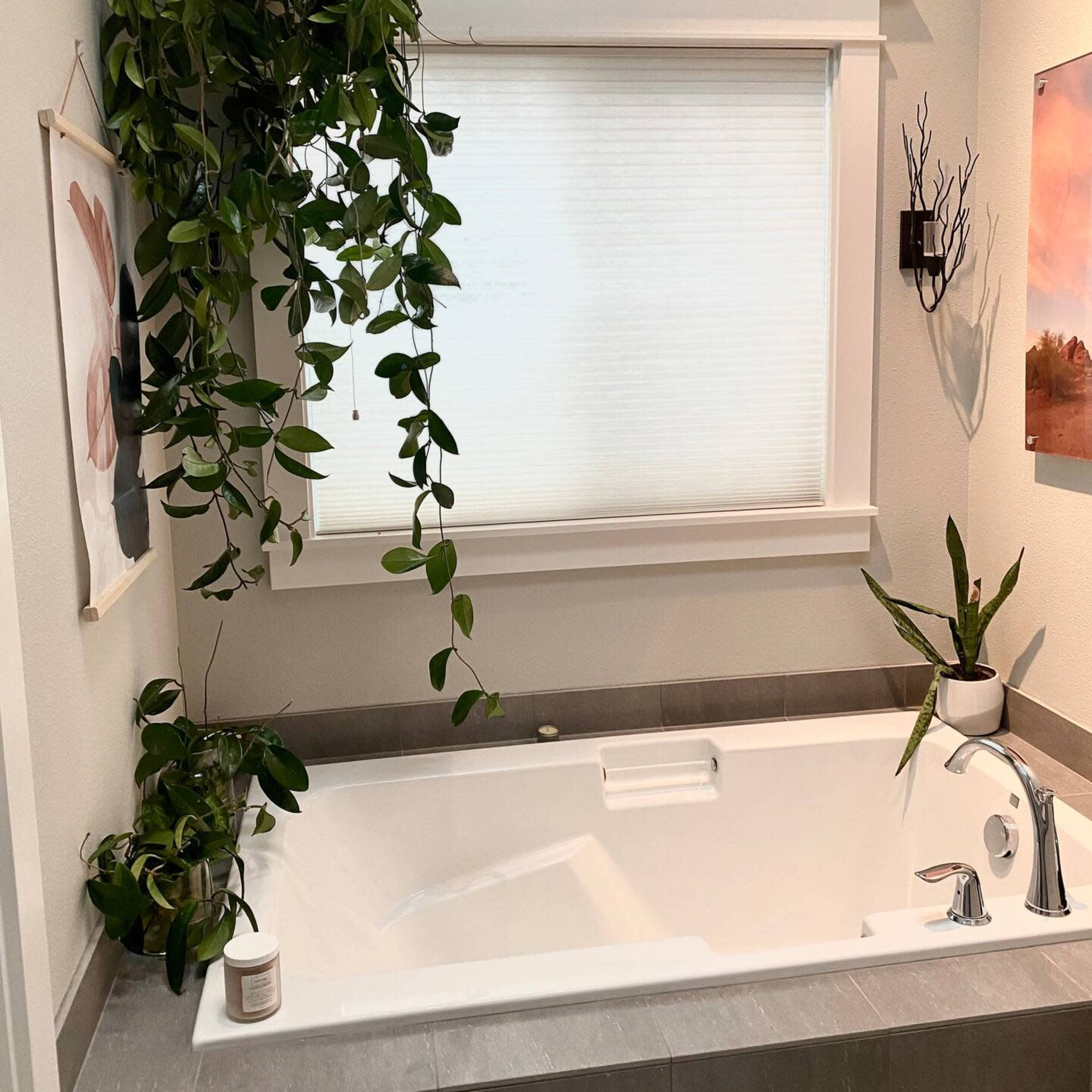 A bathroom with a white bathtub, plants positioned around the tub, and a window covered by a sheer blind.