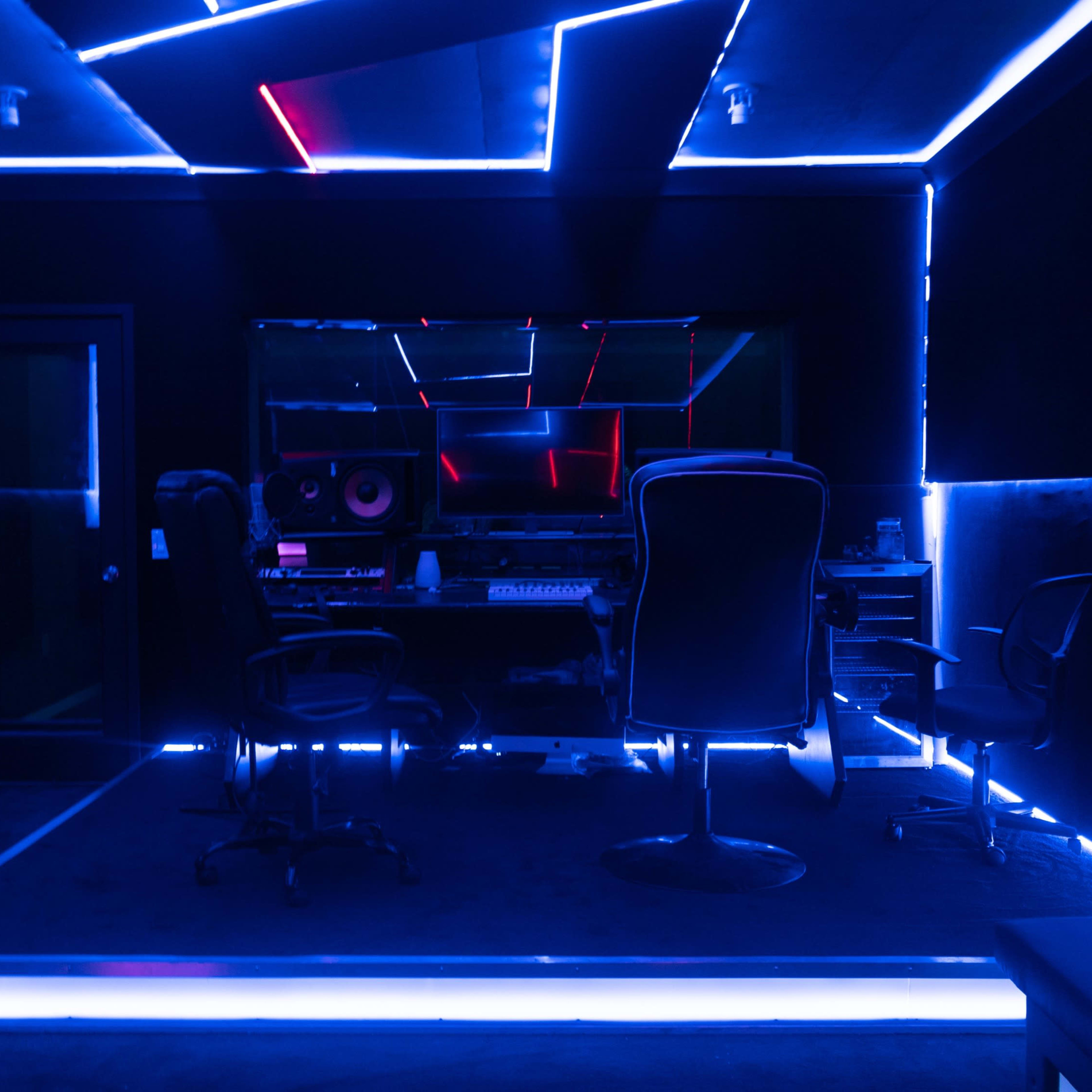 A music studio with blue LED lighting, featuring a control desk and several chairs.