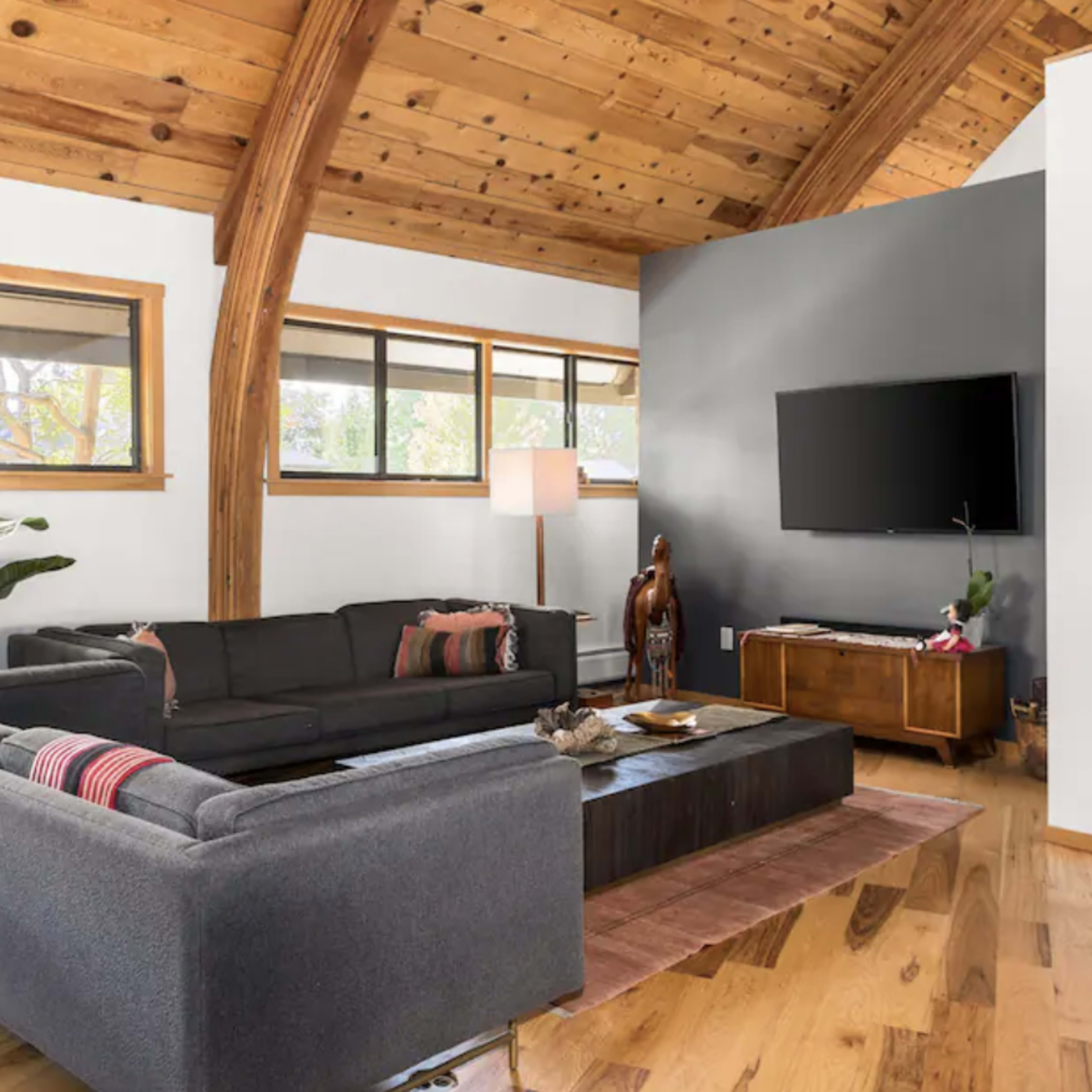A modern living room featuring a dark sectional sofa, a central coffee table, wooden ceilings, and a mounted television on a gray wall.