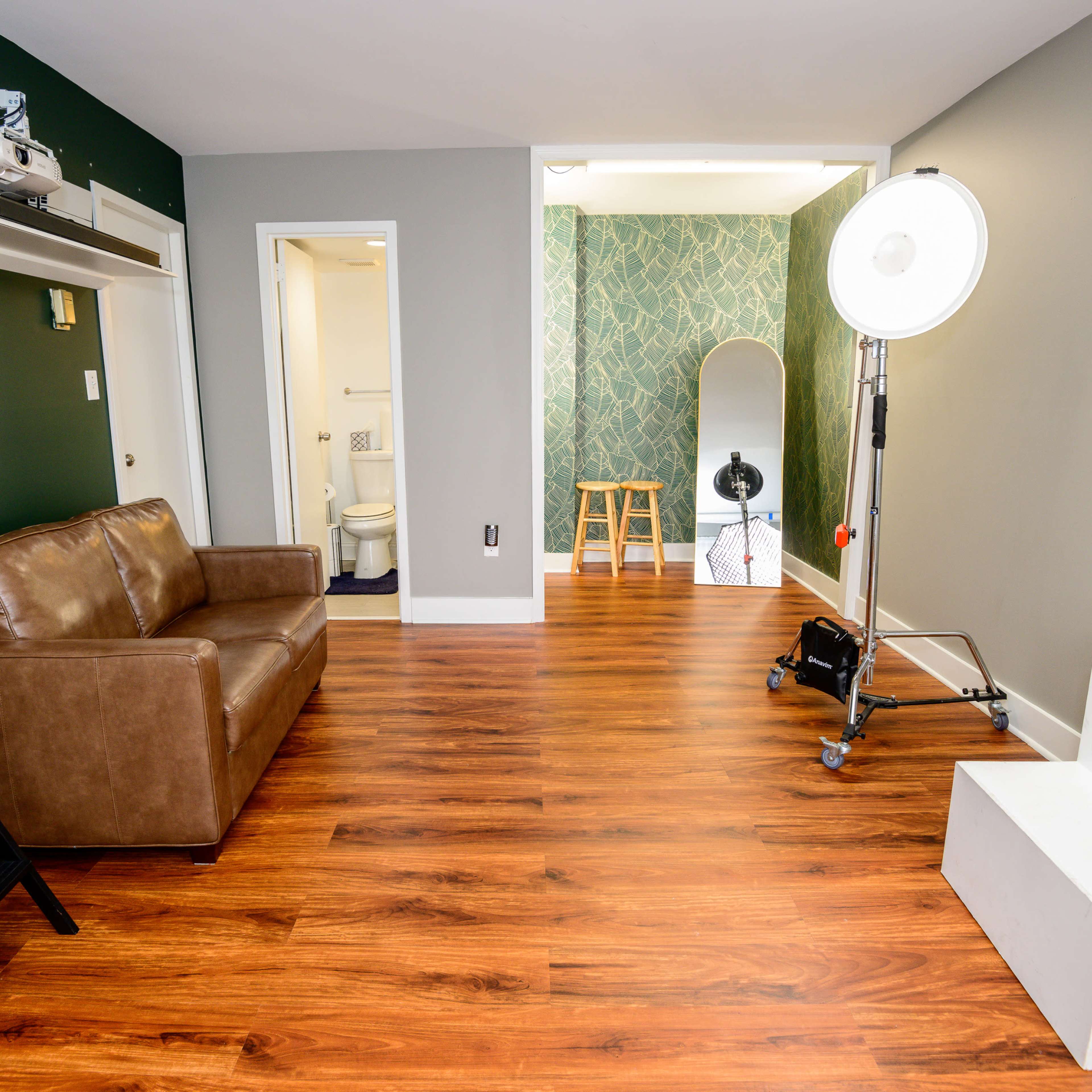 An indoor space featuring a brown sofa, a projector on a shelf, a green accent wall, wooden flooring, and a small staircase leading to a well-lit area with a mirror and stools.