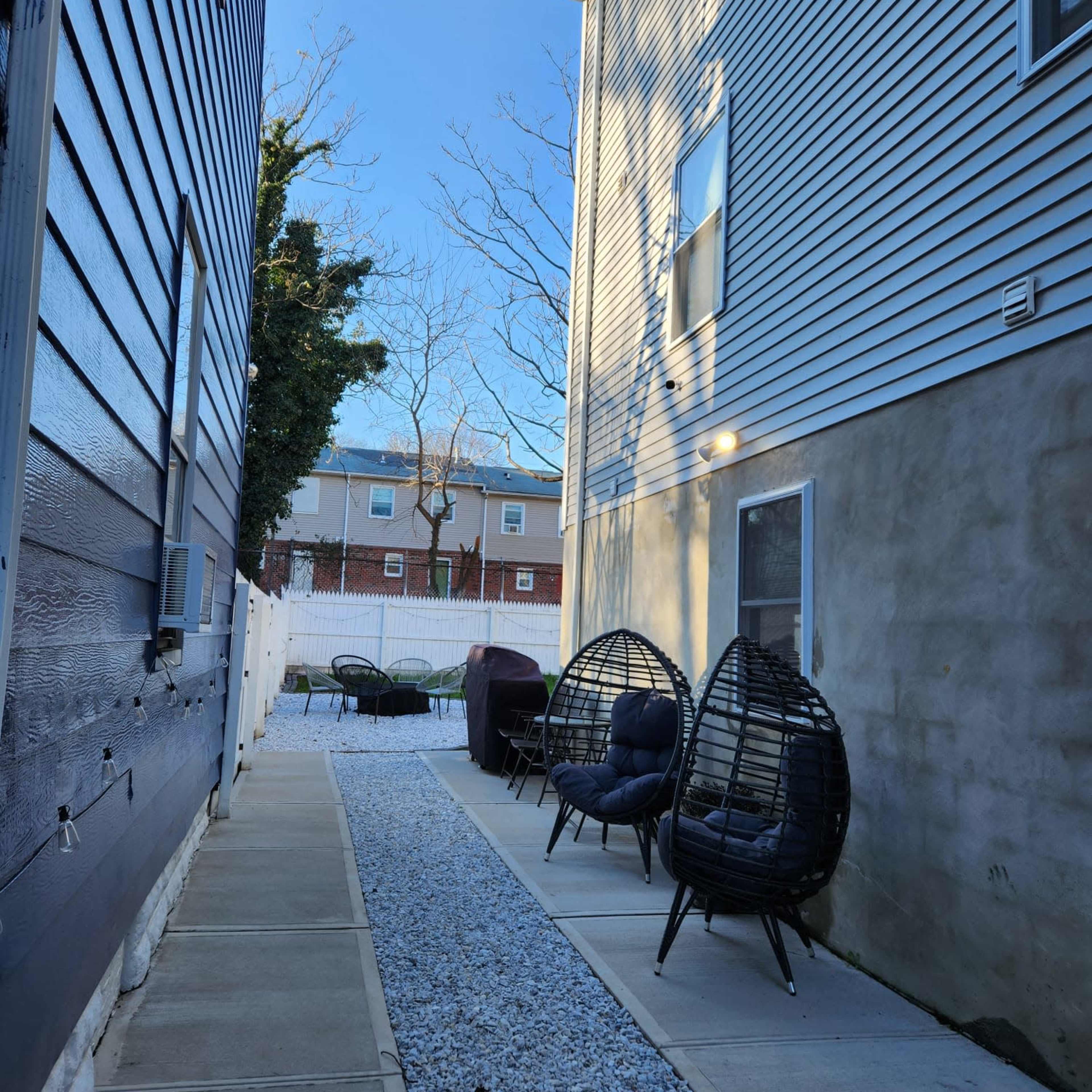 A narrow path lined with gravel leads to a cozy seating area featuring two rattan chairs beside a building.