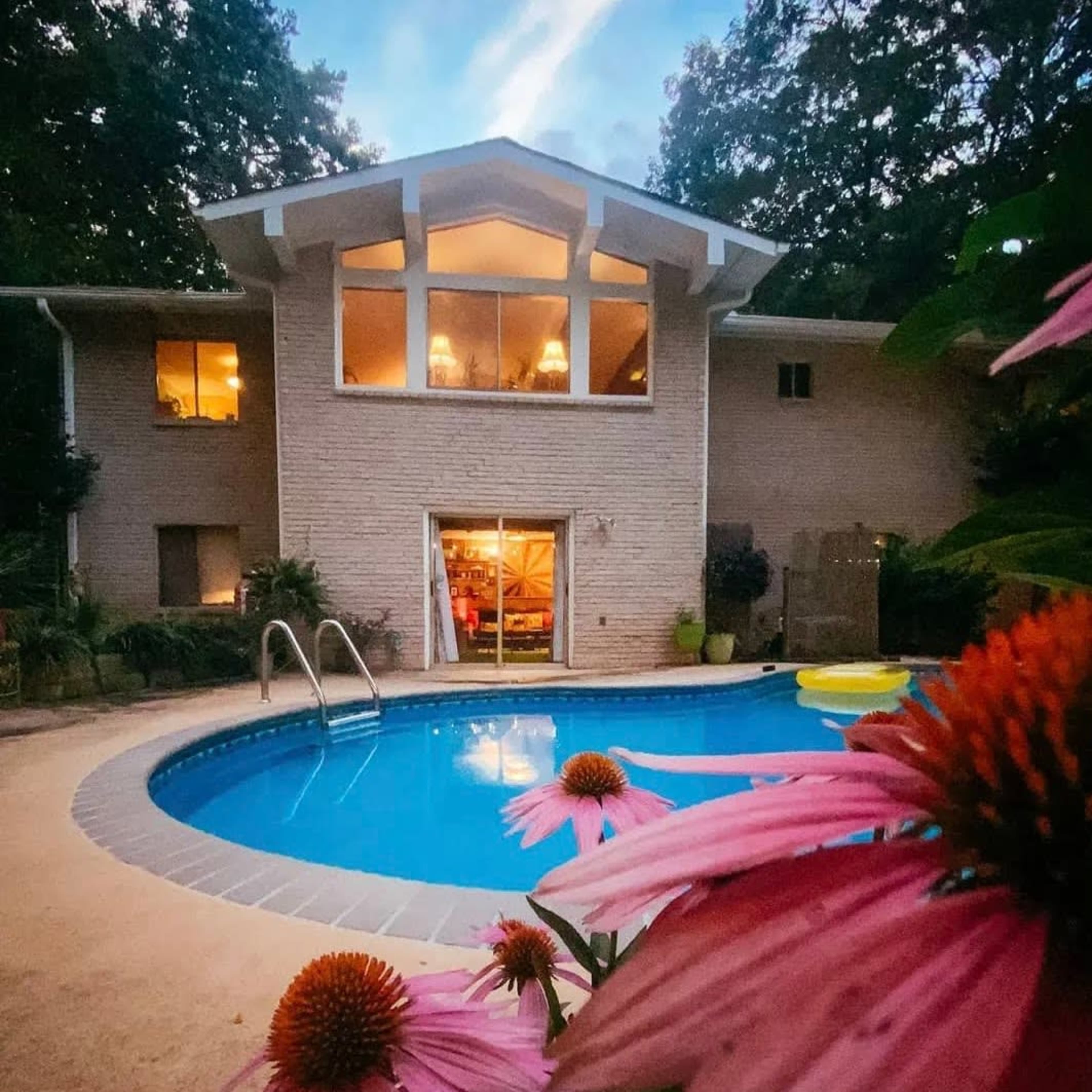 A backyard with a swimming pool in front of a two-story house, framed by flowering plants, under a twilight sky.