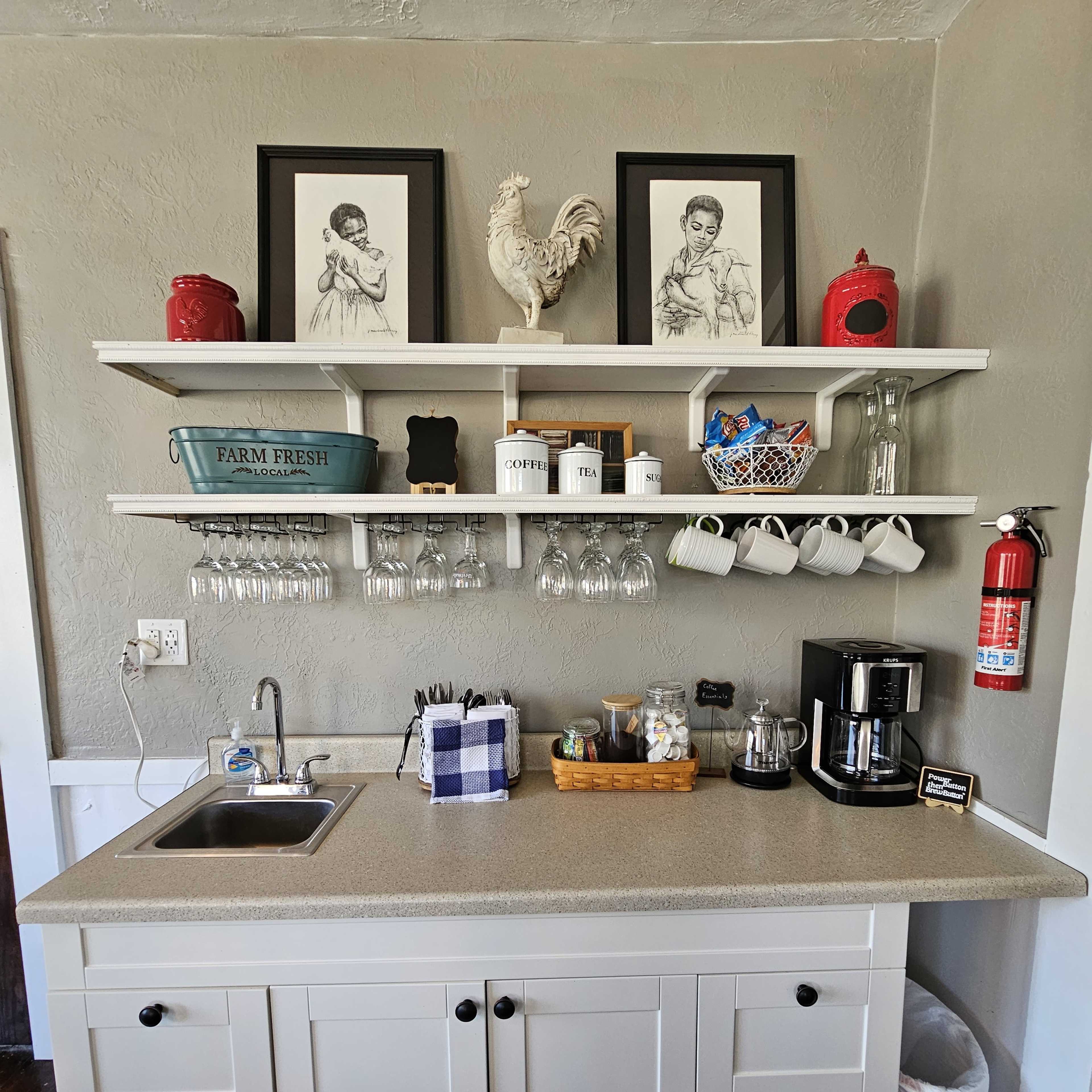 A simple kitchen area featuring a countertop with a sink, coffee maker, and storage for mugs, alongside a shelf displaying framed artwork and a rooster statue.