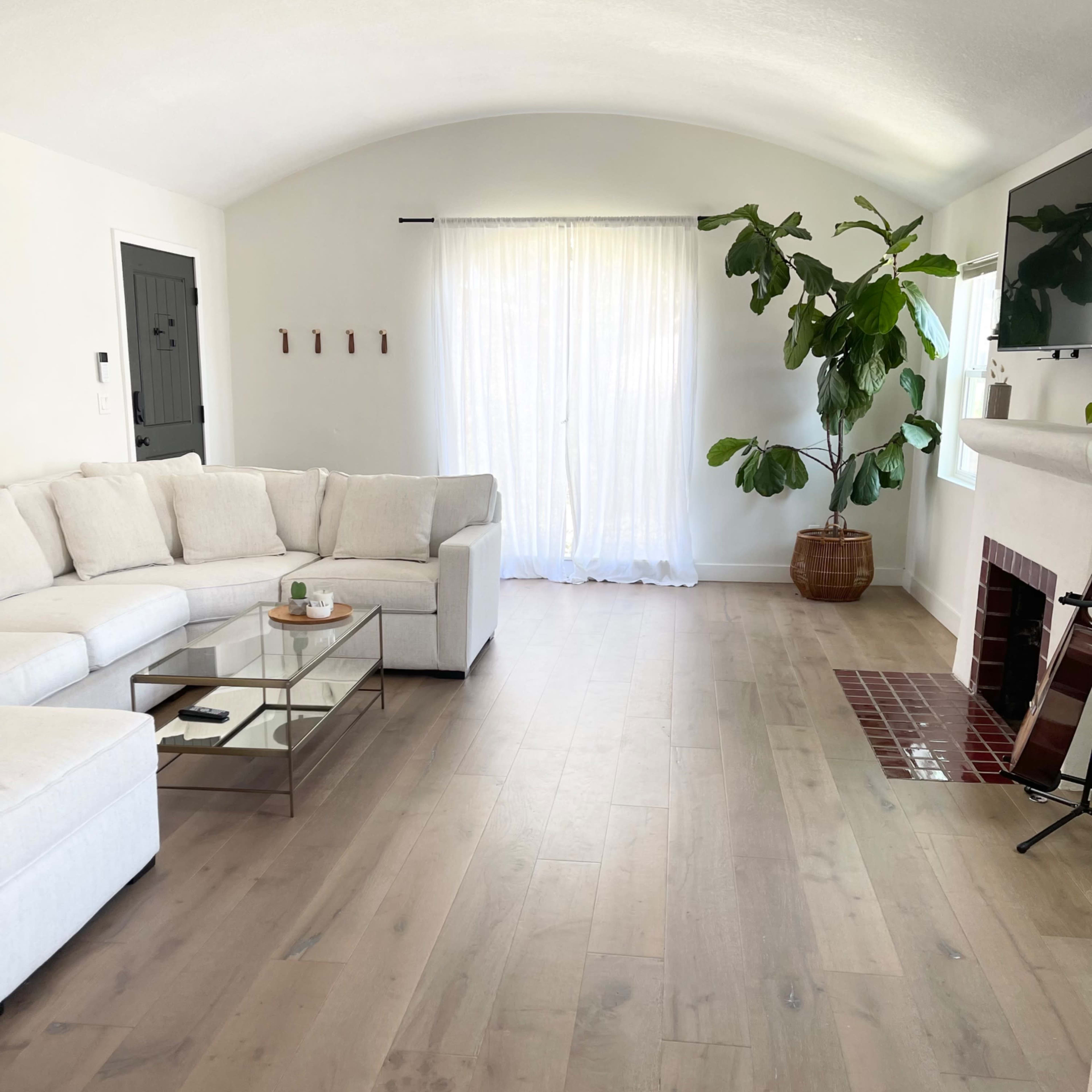 A spacious living room with a white sectional sofa, a glass coffee table, a large window covered with sheer curtains, and a potted plant in the corner.