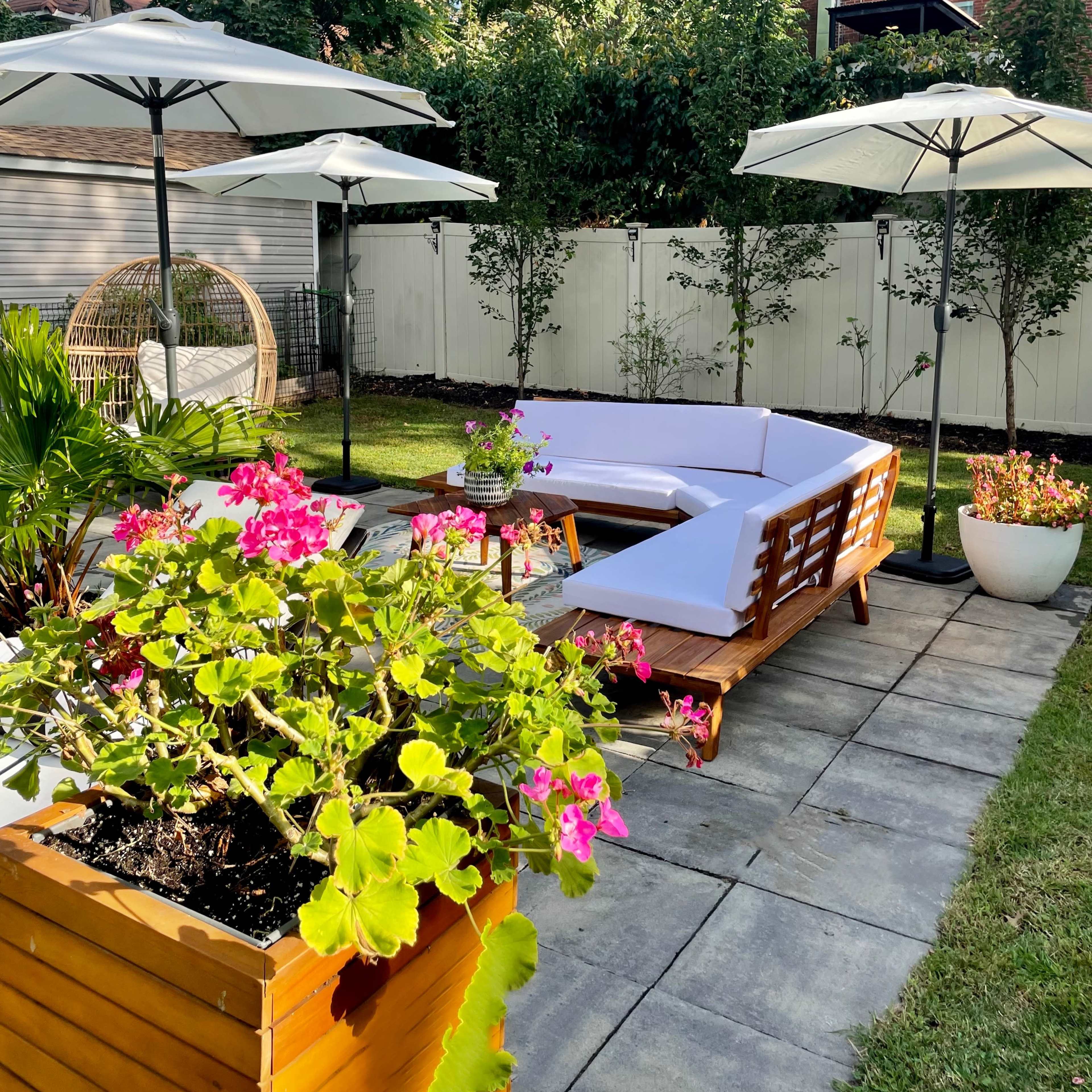 A backyard space with a white sectional sofa, several umbrellas, planters filled with colorful flowers, and a wicker chair under a tree.