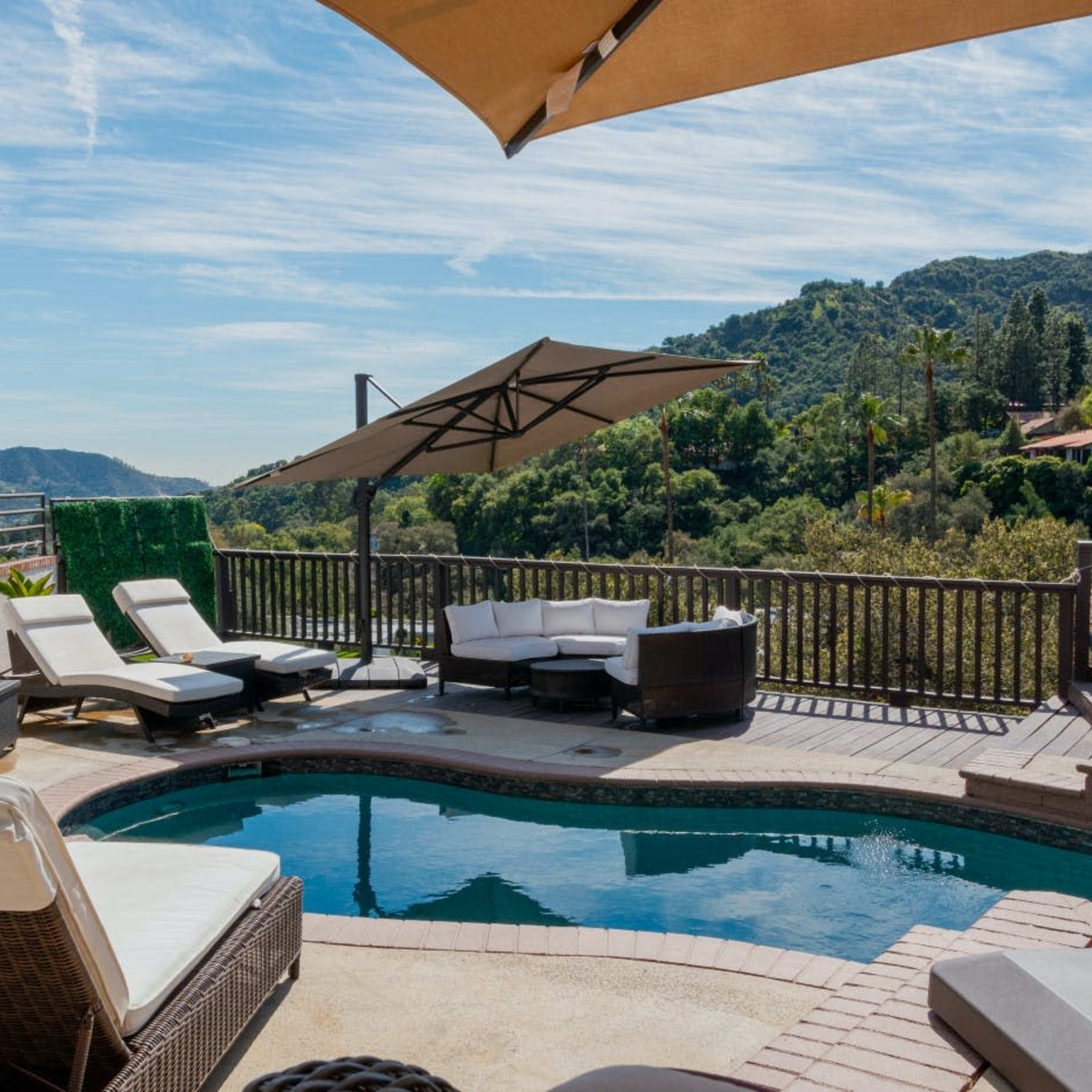 A poolside area features lounge chairs and an umbrella, overlooking a mountainous landscape.