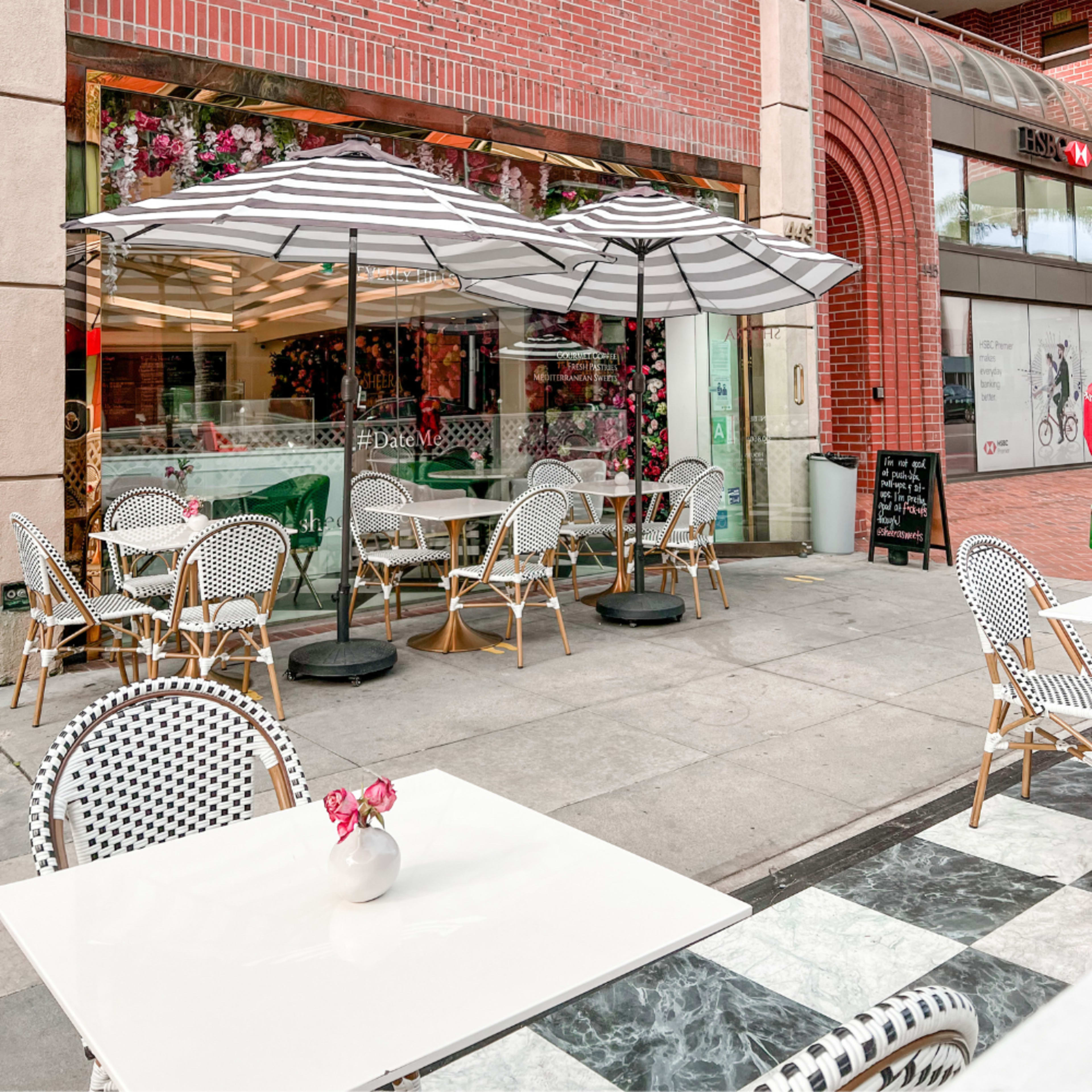 A sidewalk café features striped umbrellas, outdoor tables, and floral decorations in its window.