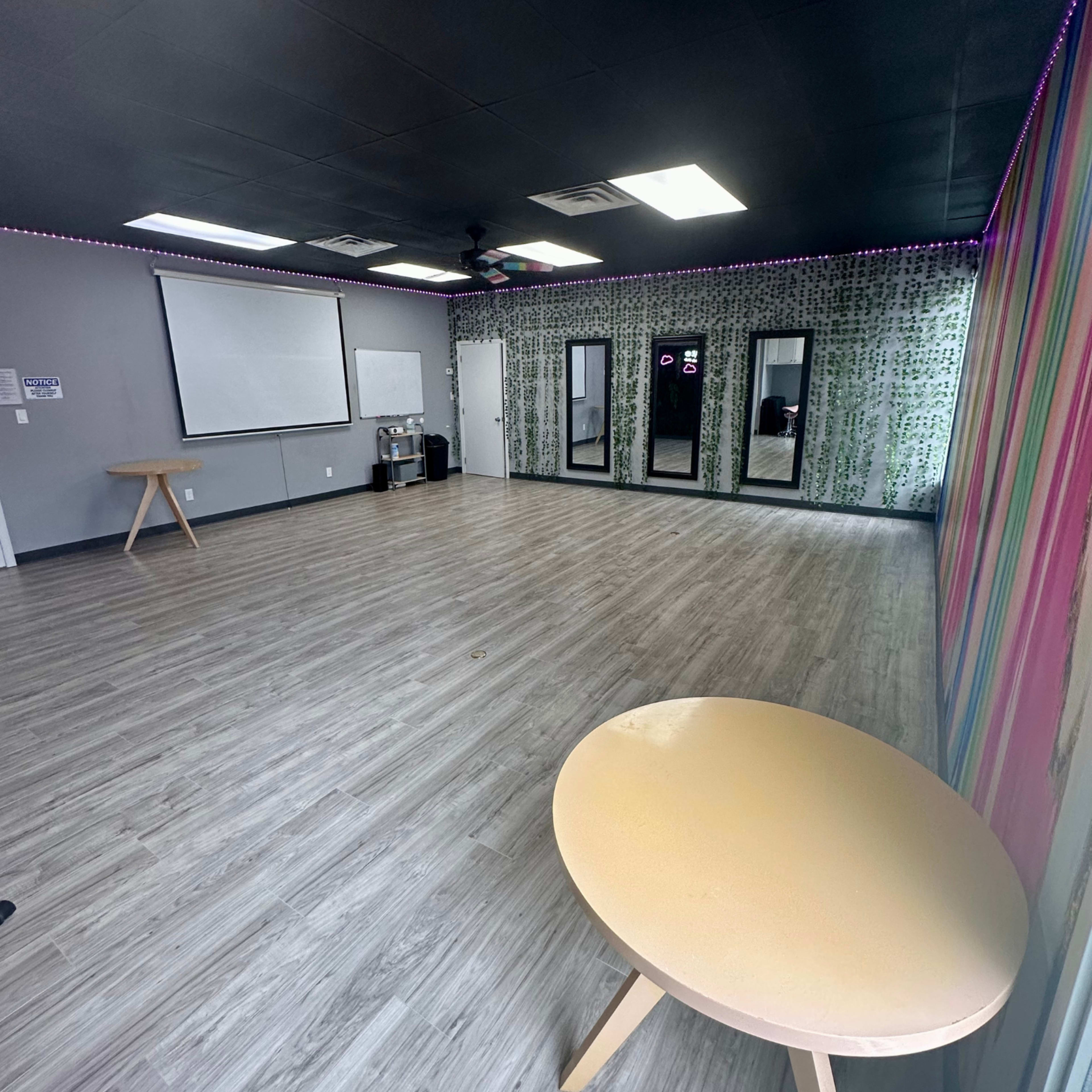 A spacious, empty room features a light-colored wooden floor, a striped wall, a projector screen, and several mirrors along one side.