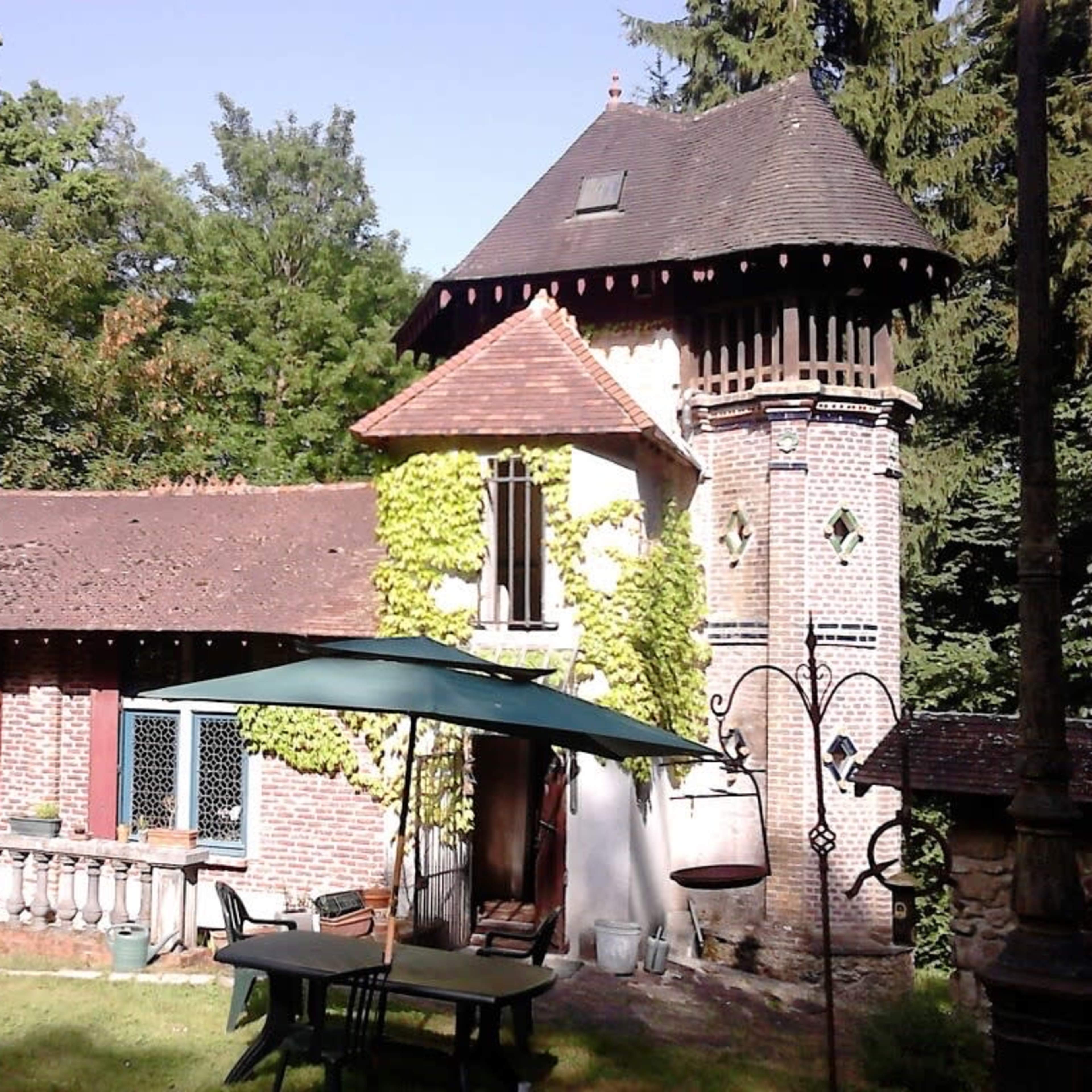 A brick house with a tower is surrounded by trees and features a green lawn and outdoor furniture.