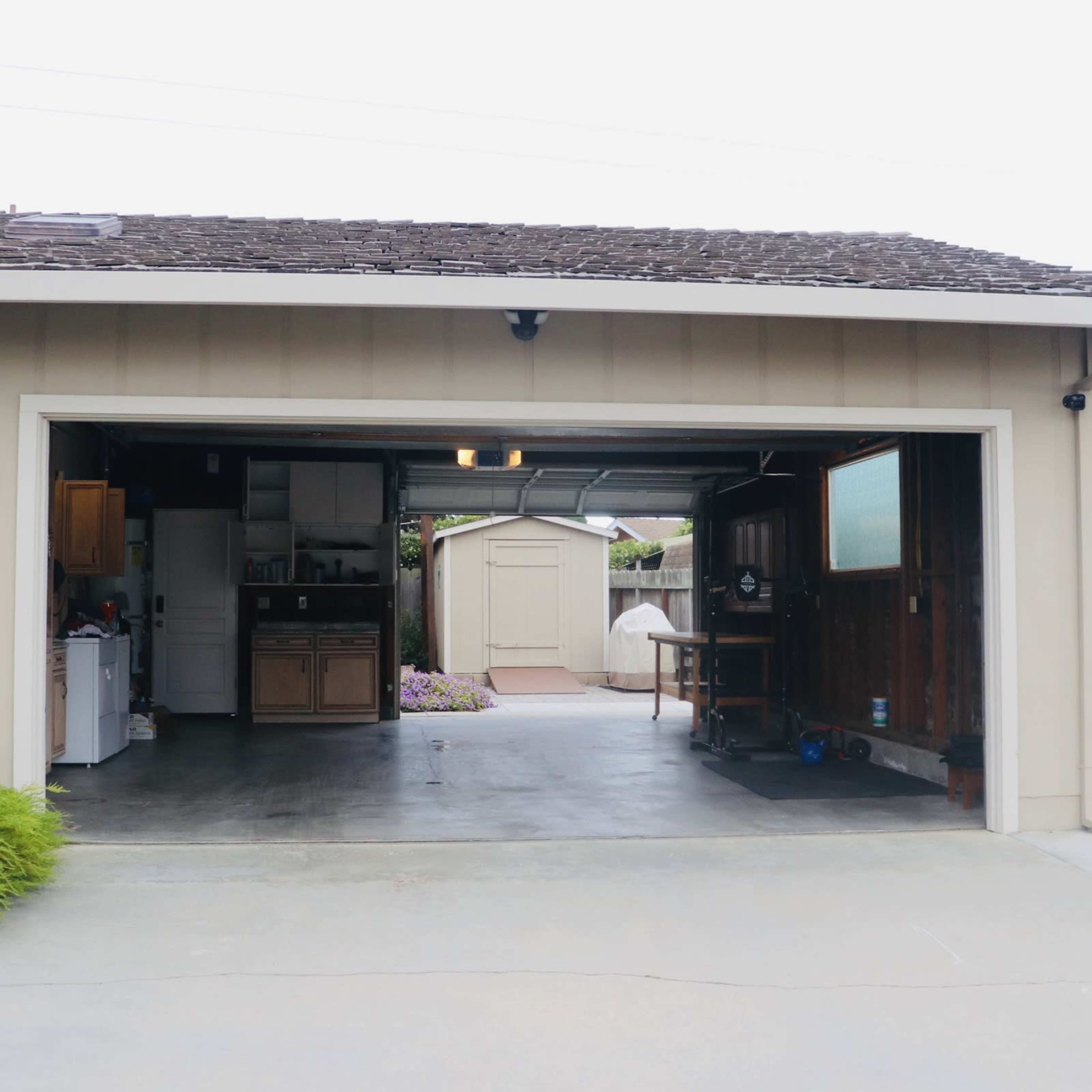 A garage with an open door reveals a kitchen area to the left and leads to a backyard shed in the distance.