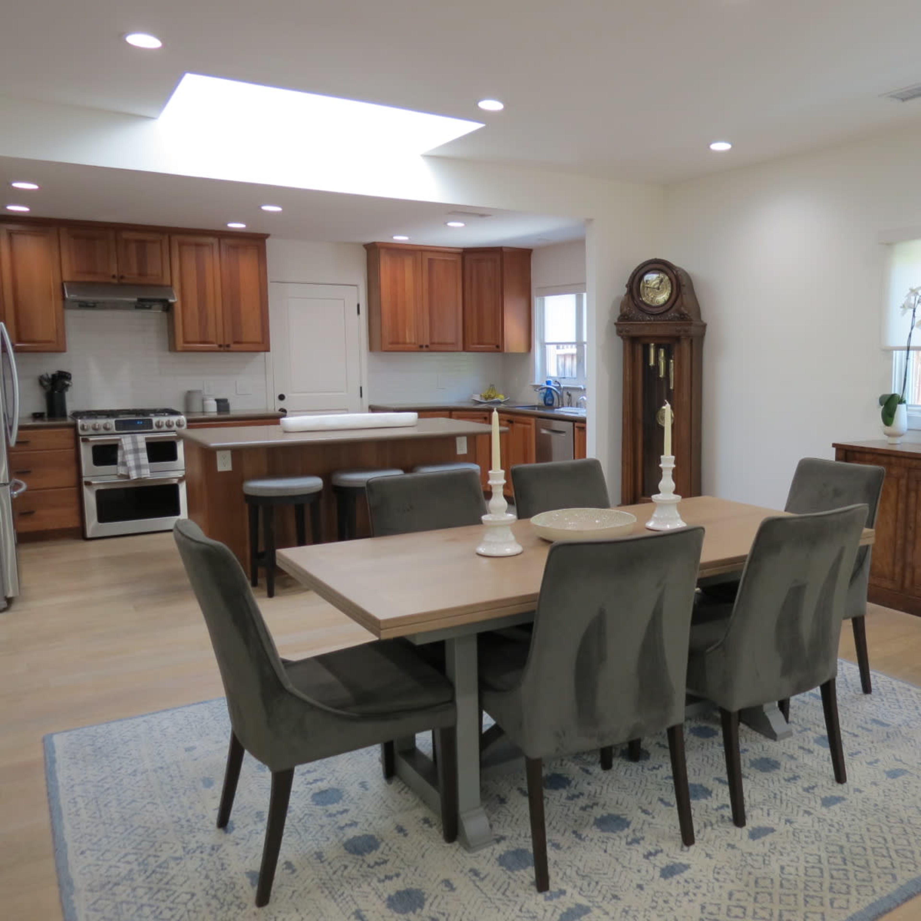 A modern dining area features a wooden table with gray chairs, surrounded by a well-lit kitchen with wooden cabinets and a clock against the wall.