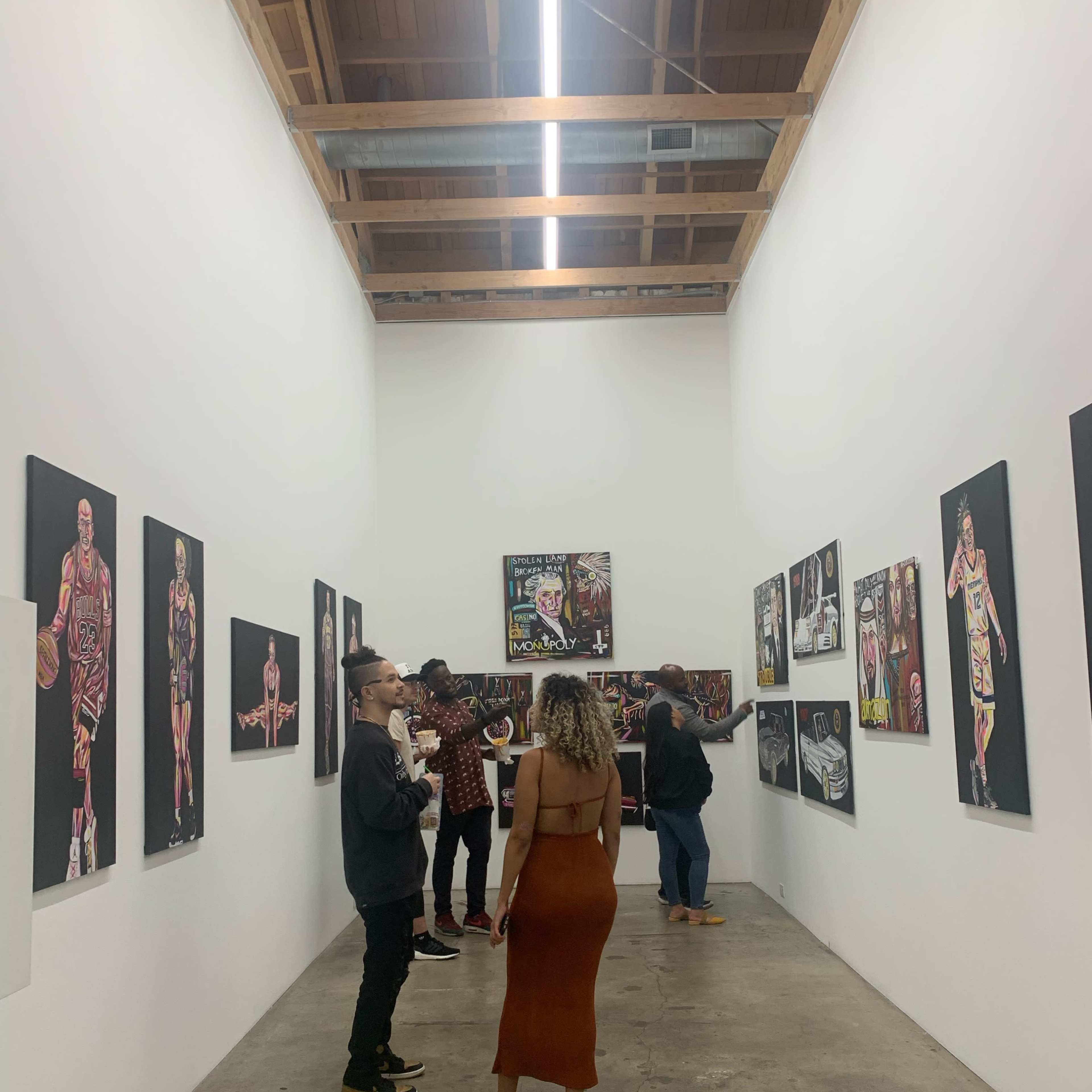 A group of people are viewing artwork in a narrow gallery space with various paintings displayed on the walls.