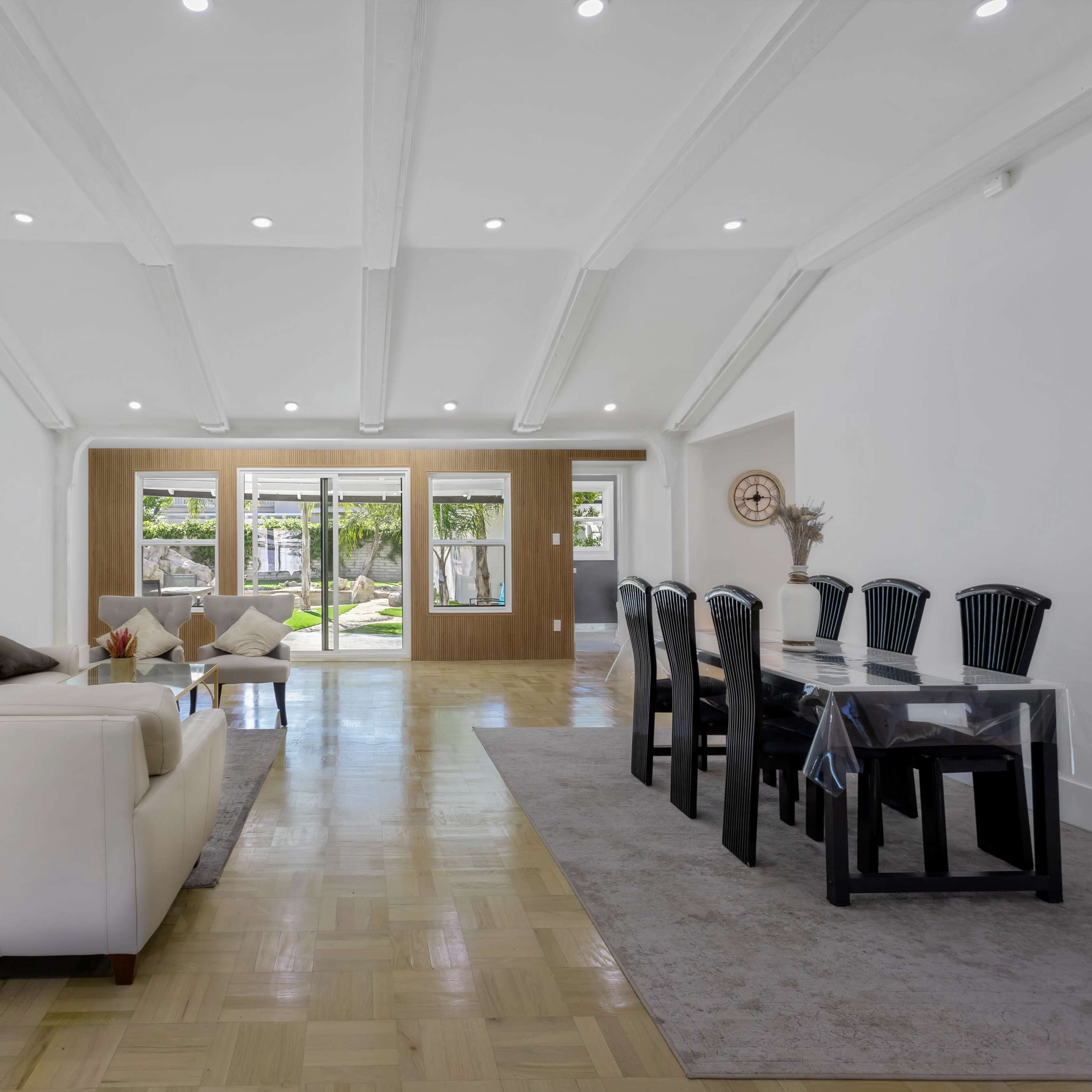 A spacious living area featuring a white sofa, a glass dining table with black chairs, and large windows that offer a view of an outdoor space.