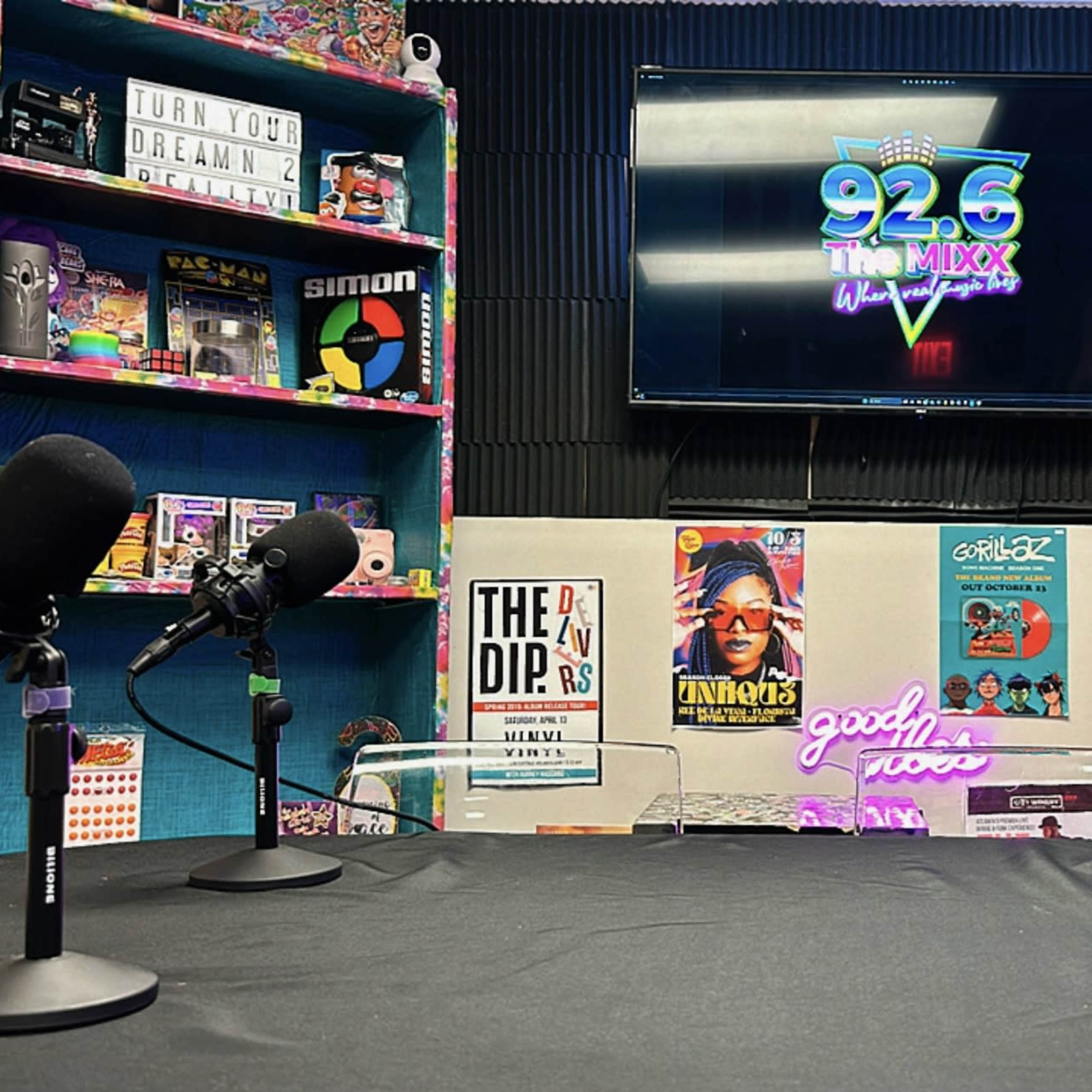 A radio studio setup with microphones arranged on a table, a television displaying the logo "92.6 The Mixx," and colorful shelves filled with vintage games and records in the background.