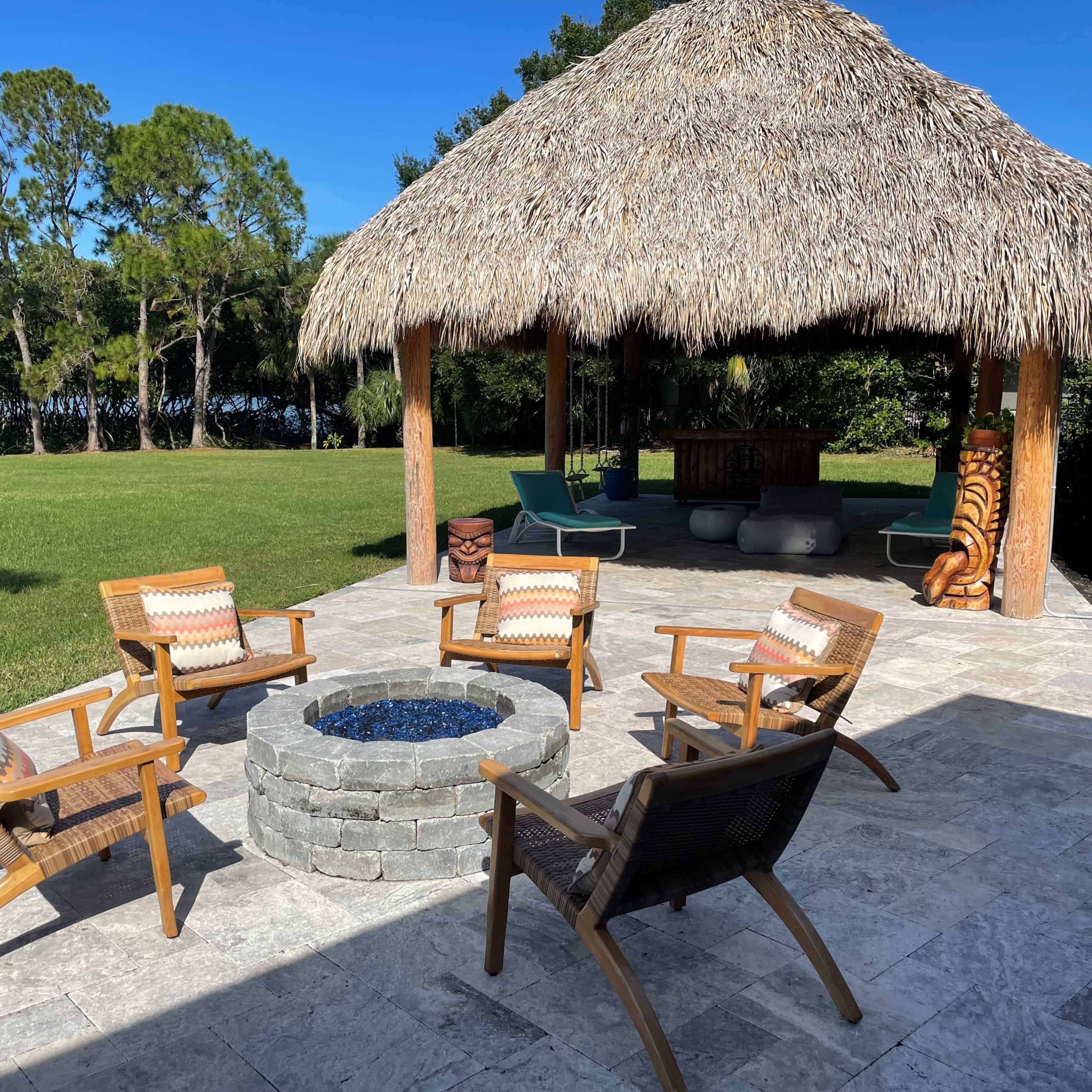 A circular stone fire pit surrounded by four wooden chairs sits beneath a thatched gazebo in a grassy outdoor space.