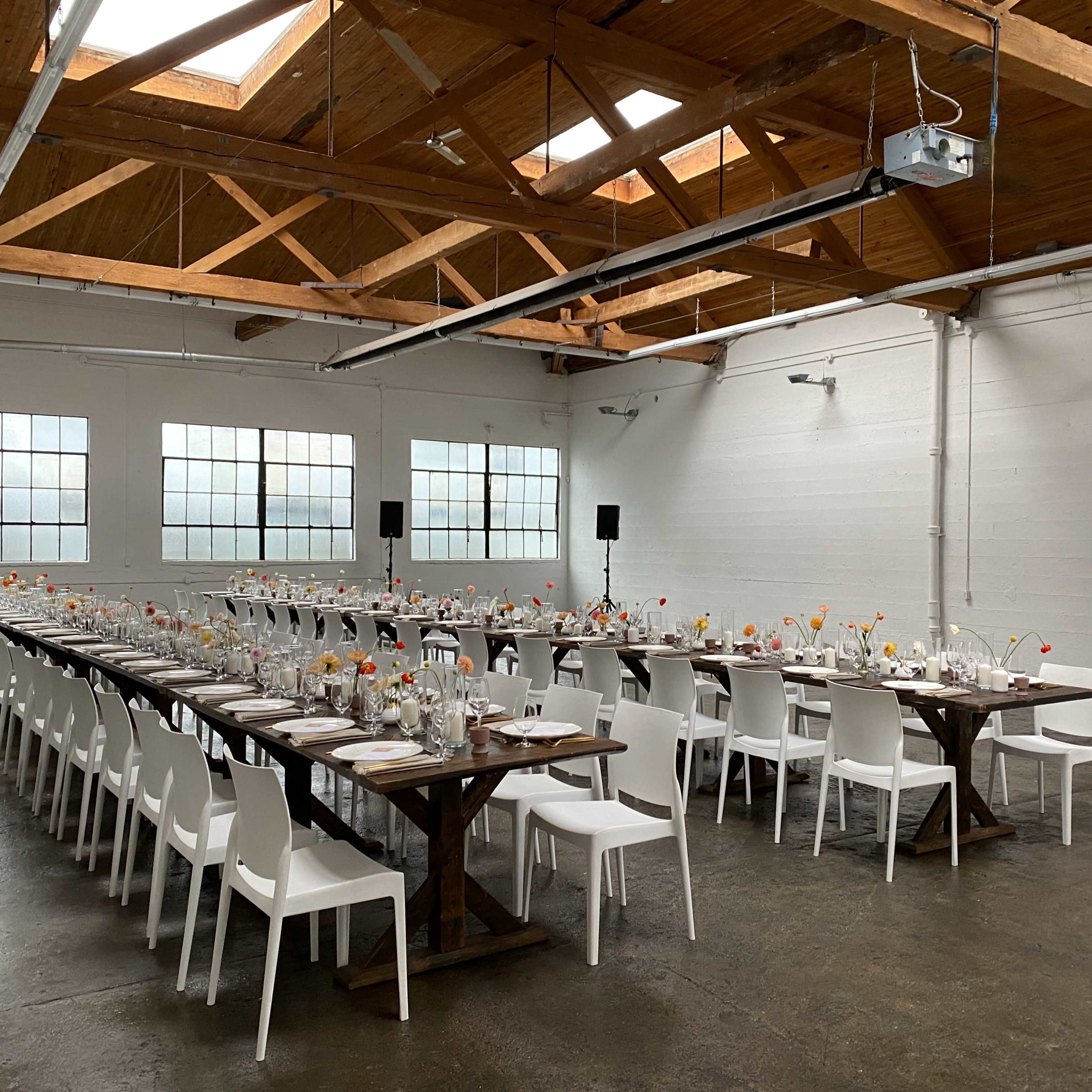 A long banquet table is set for a formal event in a spacious, well-lit room with wooden beams and large windows.