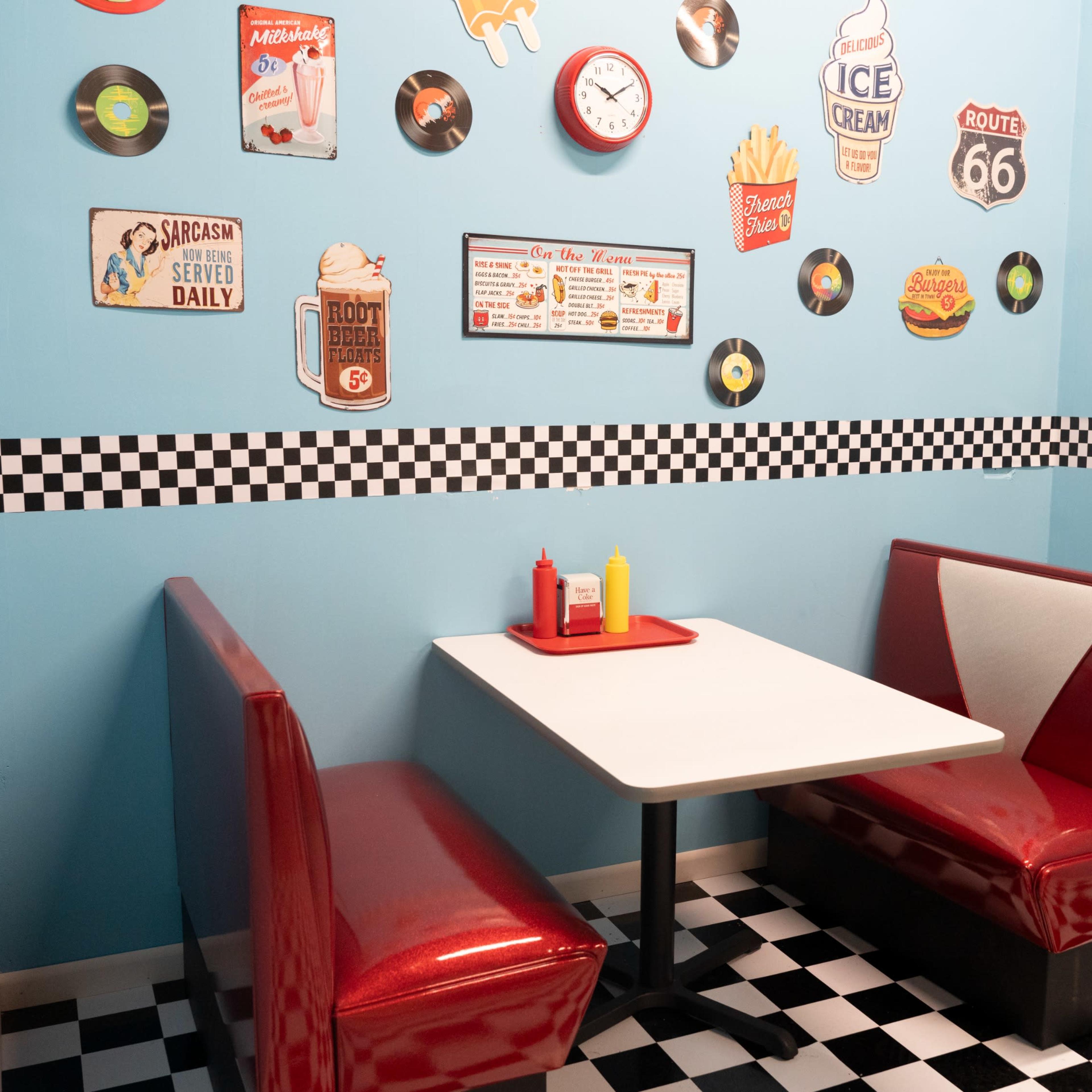 A retro diner booth with red seating, a white table, and walls decorated with vintage signs and records in a checkered pattern.
