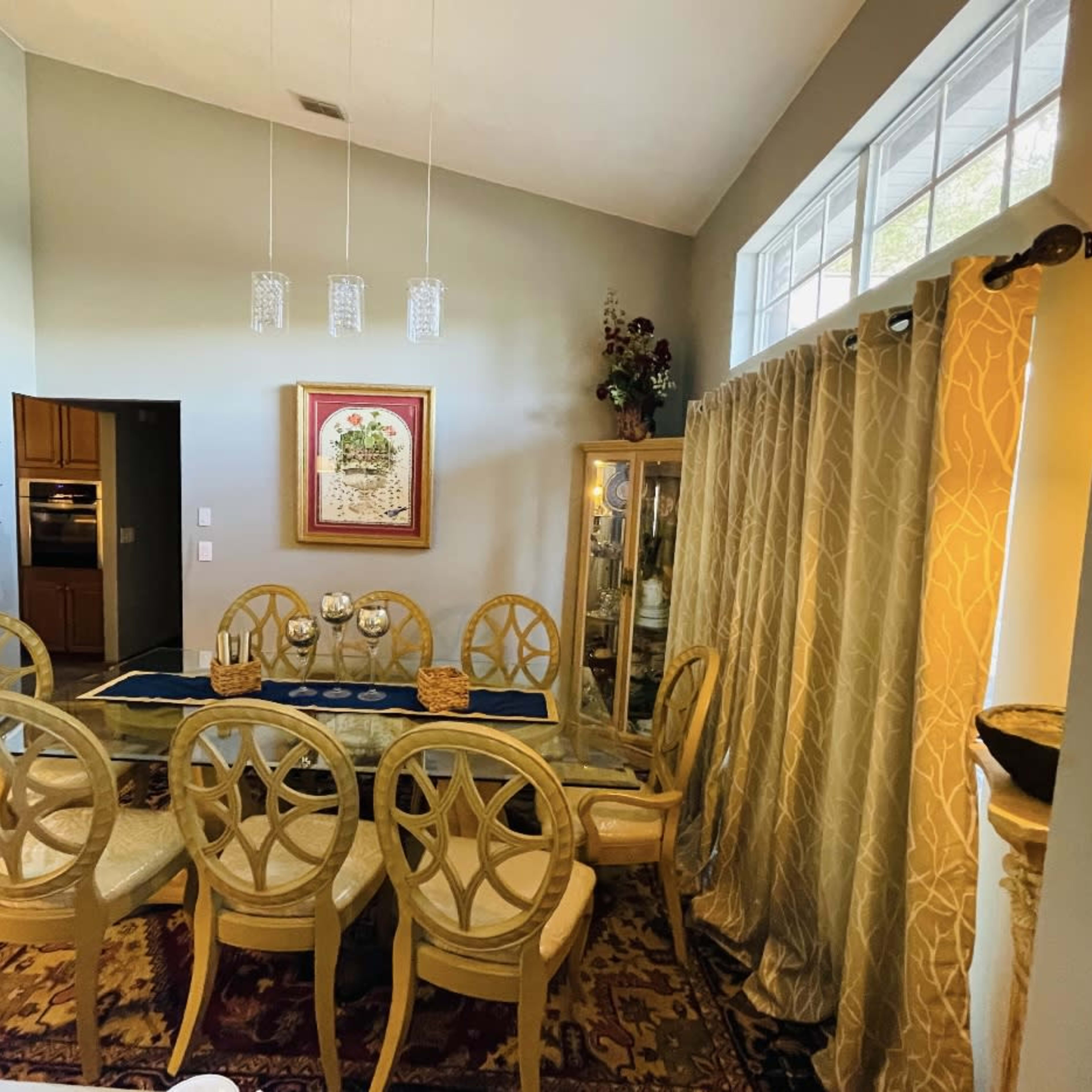 A dining area features a large table surrounded by eight chairs, with a nearby window and a decorative cabinet against the wall.