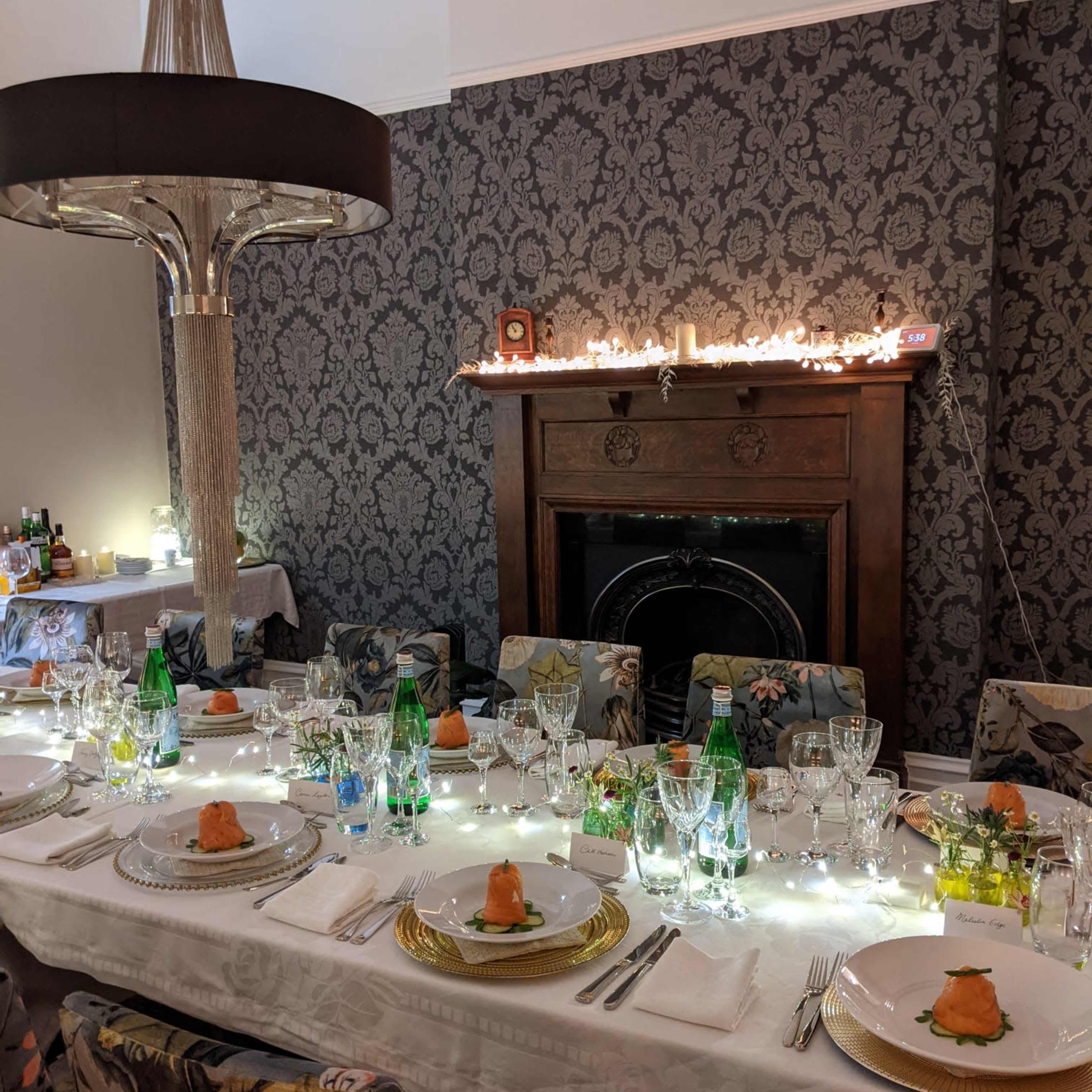 A long, elegantly set dining table with white tablecloth, gold-rimmed plates, and soft lighting in a patterned room.