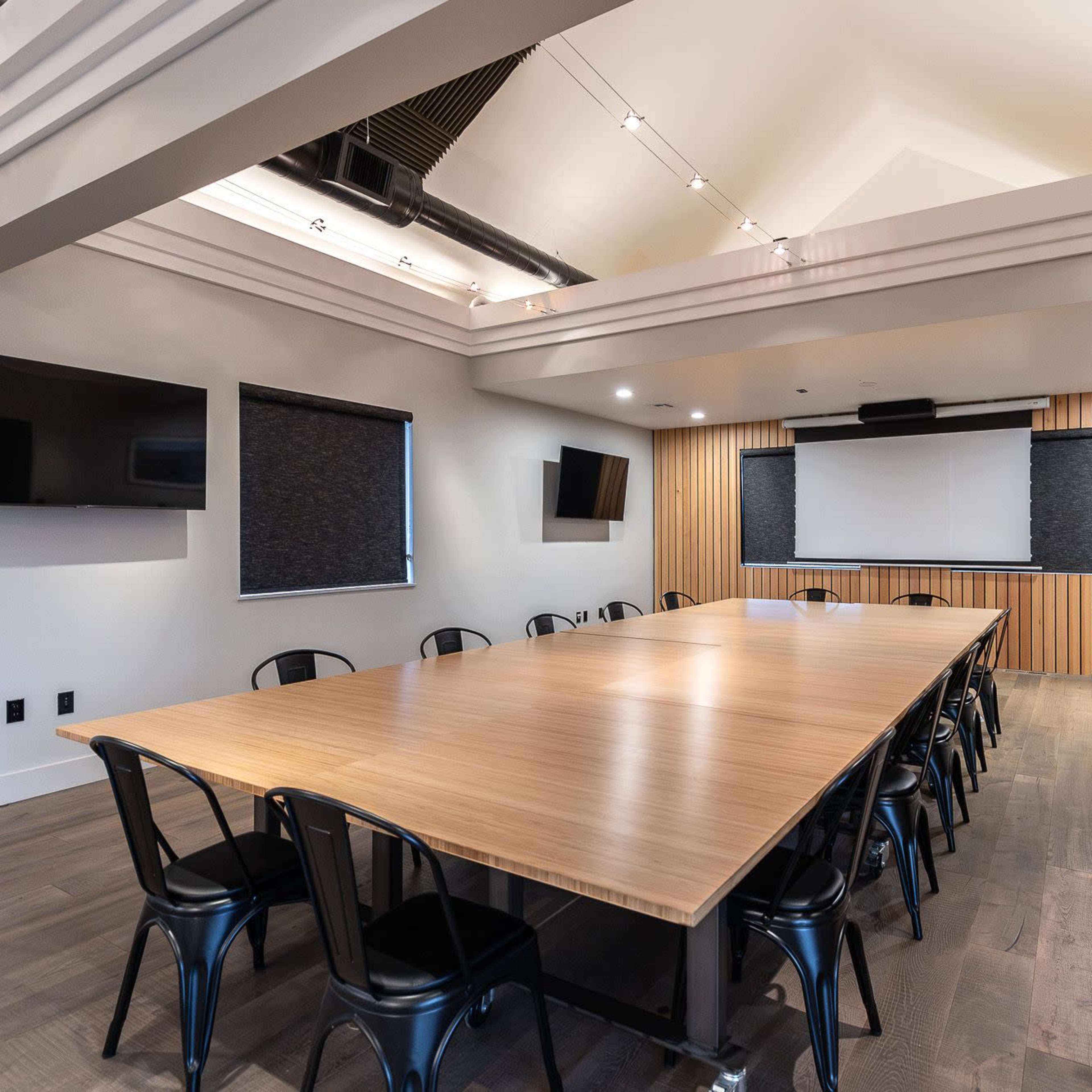 A spacious conference room with a large wooden table, black chairs, and a screen at the front, complemented by wall-mounted TVs.