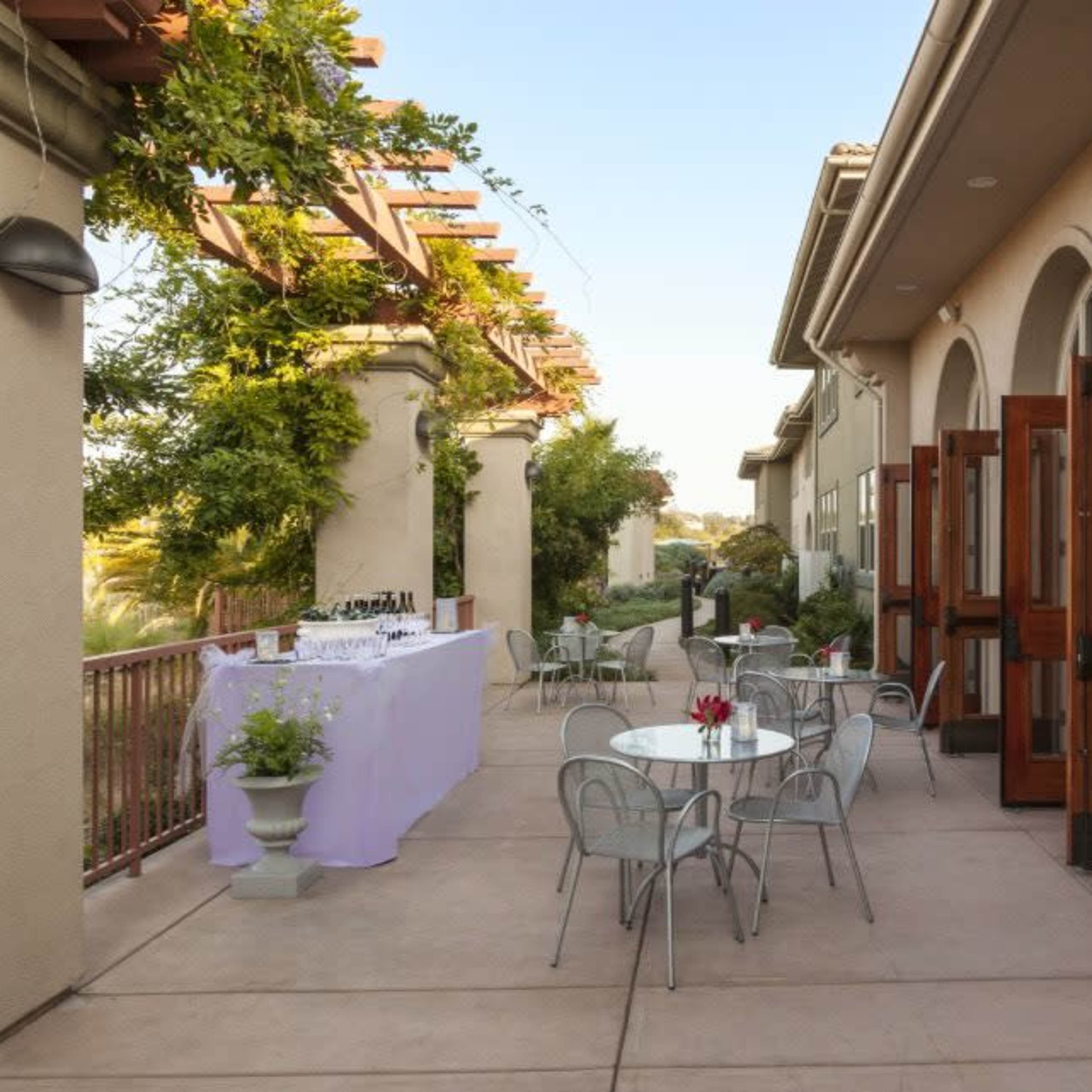 A patio area with tables and chairs, flanked by large arches and doors leading into a building, with a table set up for an event in the background.