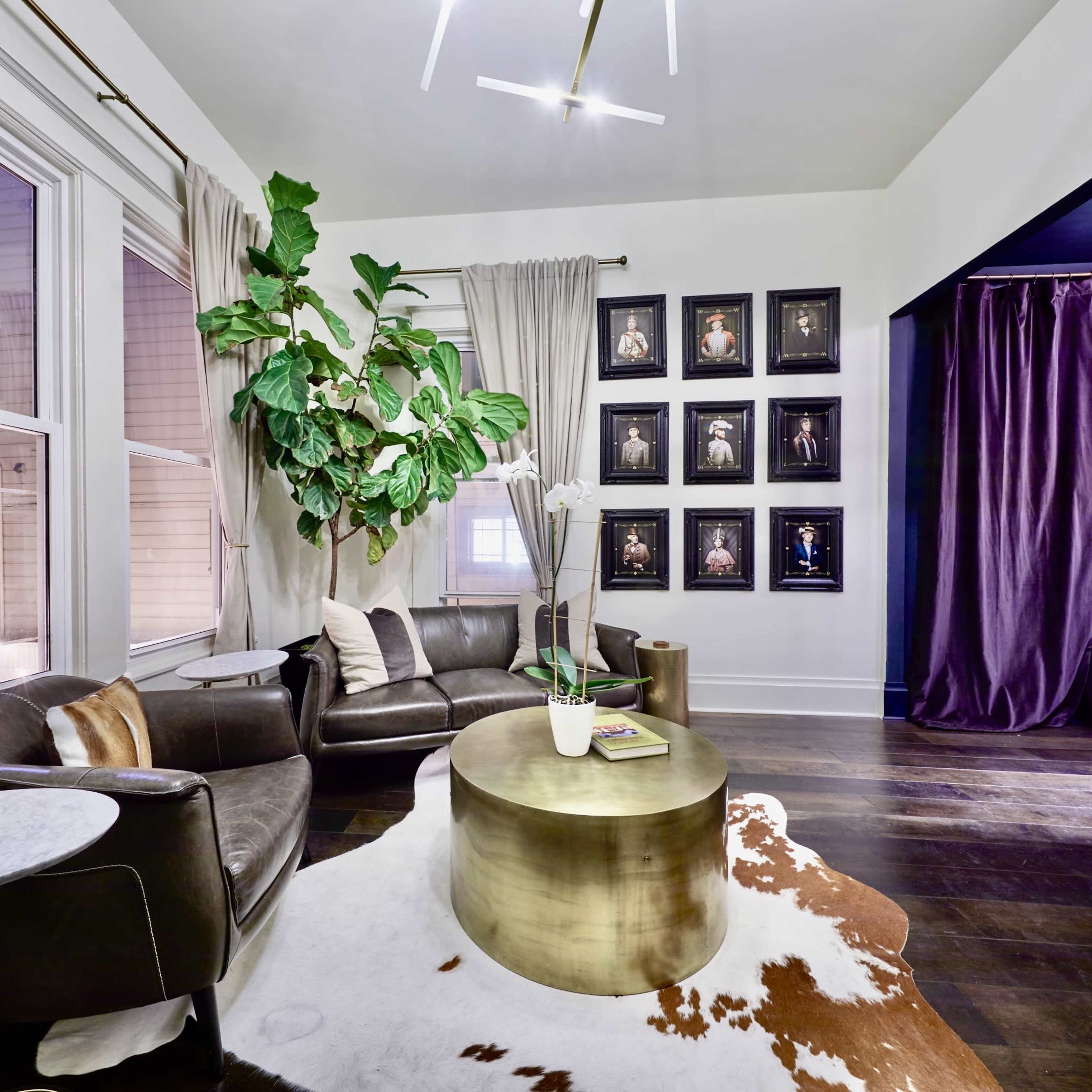 A modern living room features a dark leather sofa, a round metallic coffee table, large plants, and framed artwork on the wall.