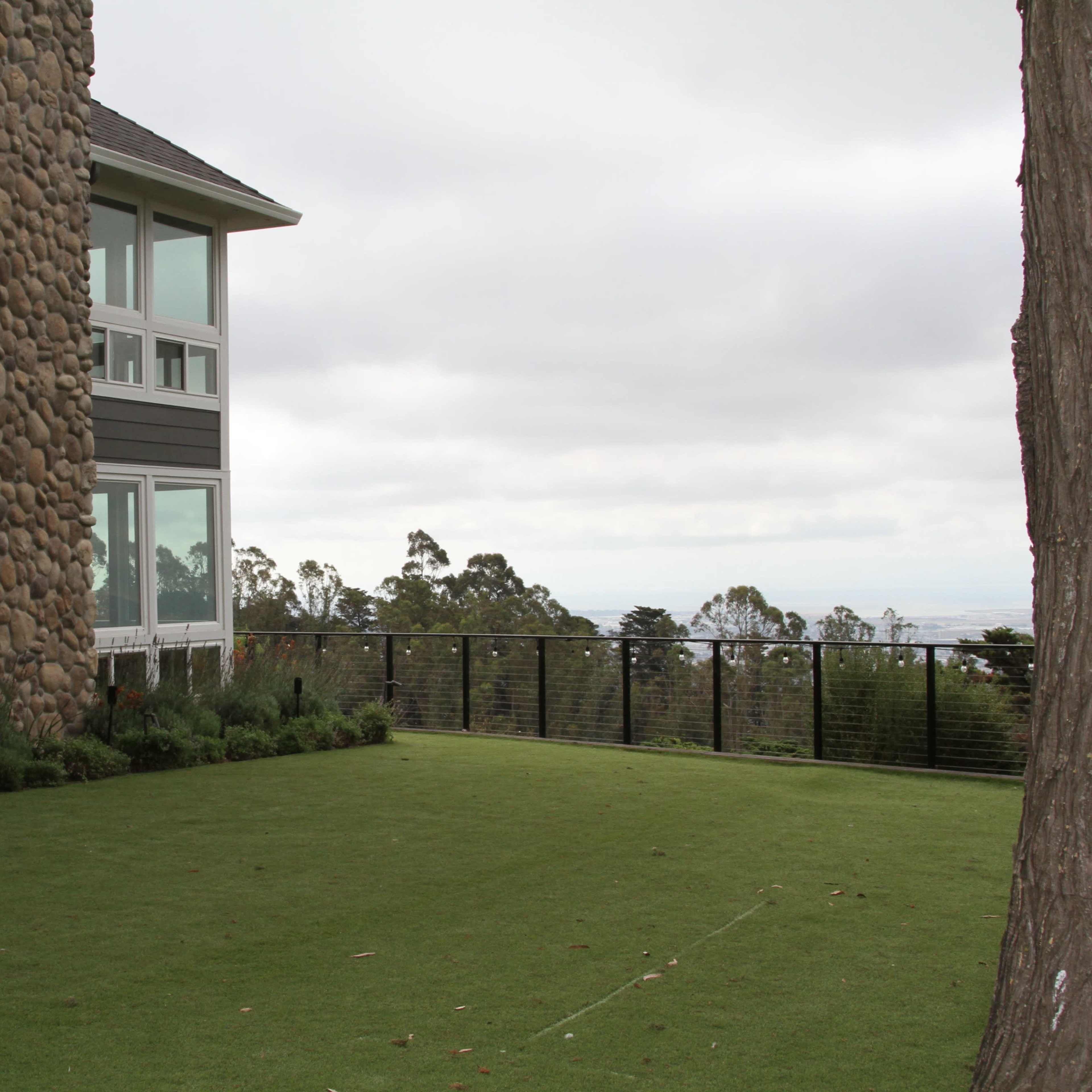 A two-story house with a stone chimney is situated beside a manicured lawn and a fence overlooking a distant landscape.