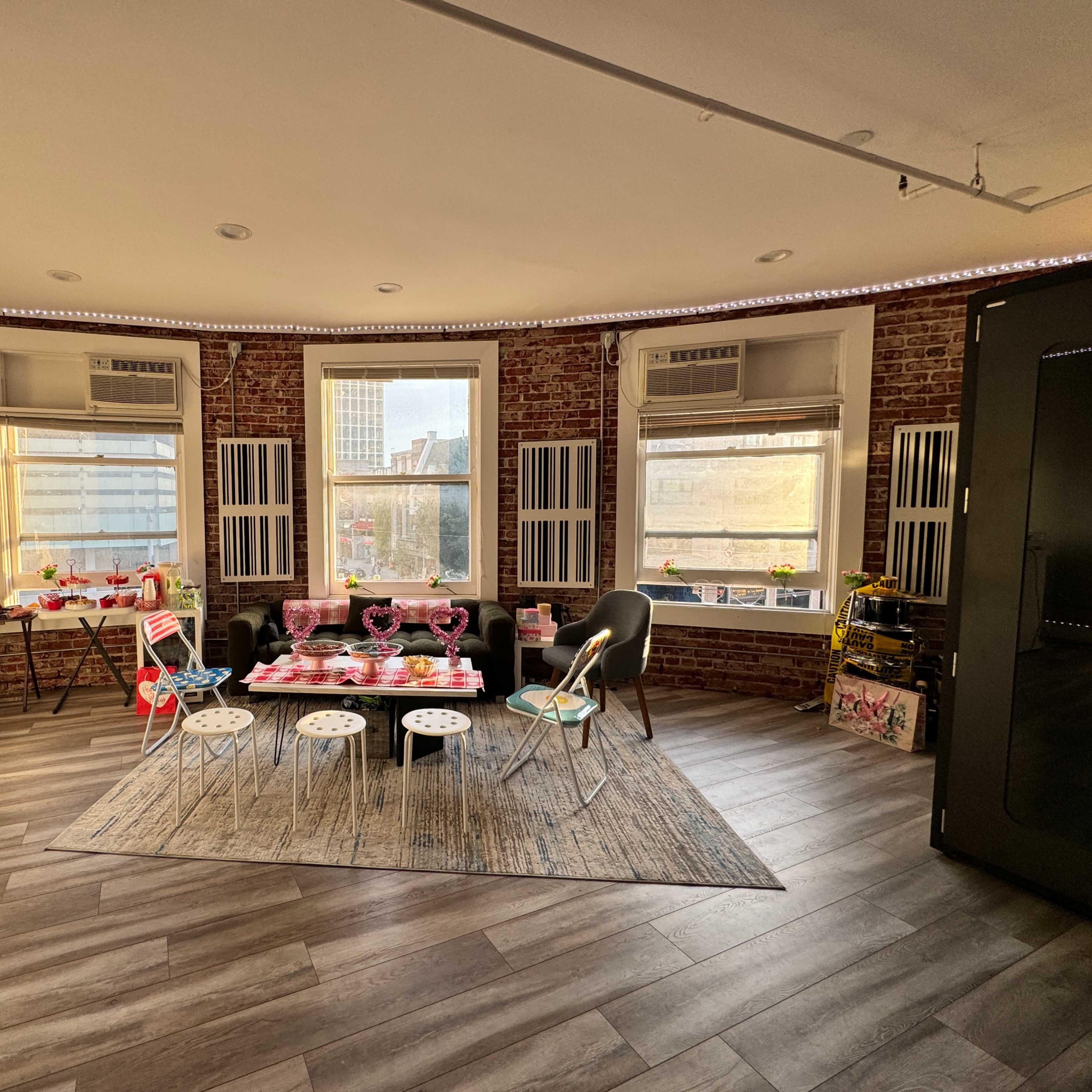The room features a round layout with exposed brick walls, three large windows, and contemporary furniture arranged around a central area with a patterned rug.