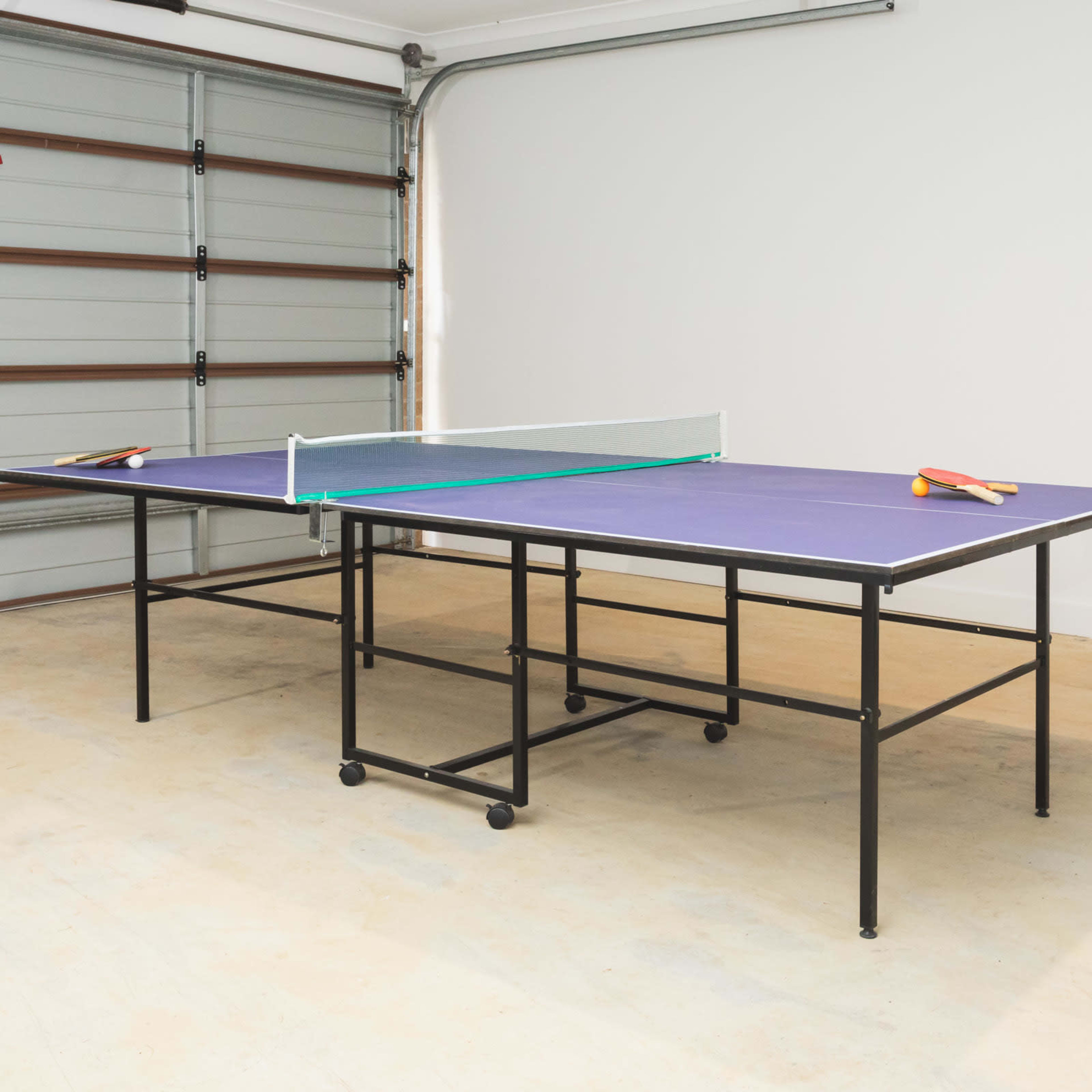 A ping pong table with paddles rests in a garage with a closed door.
