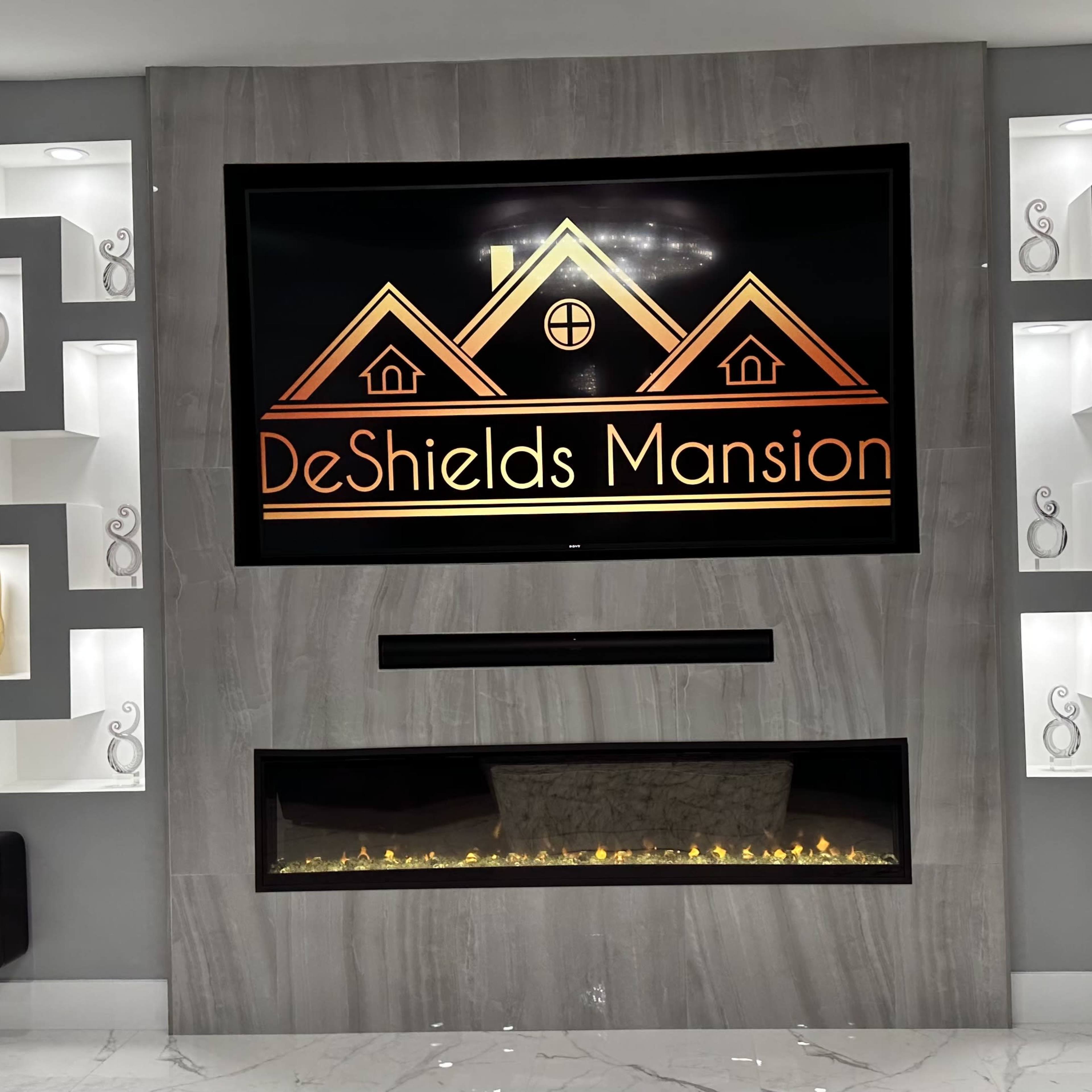 A modern living room wall featuring a large TV with the "DeShields Mansion" logo centered above a decorative fireplace, flanked by illuminated shelves displaying artistic pieces.