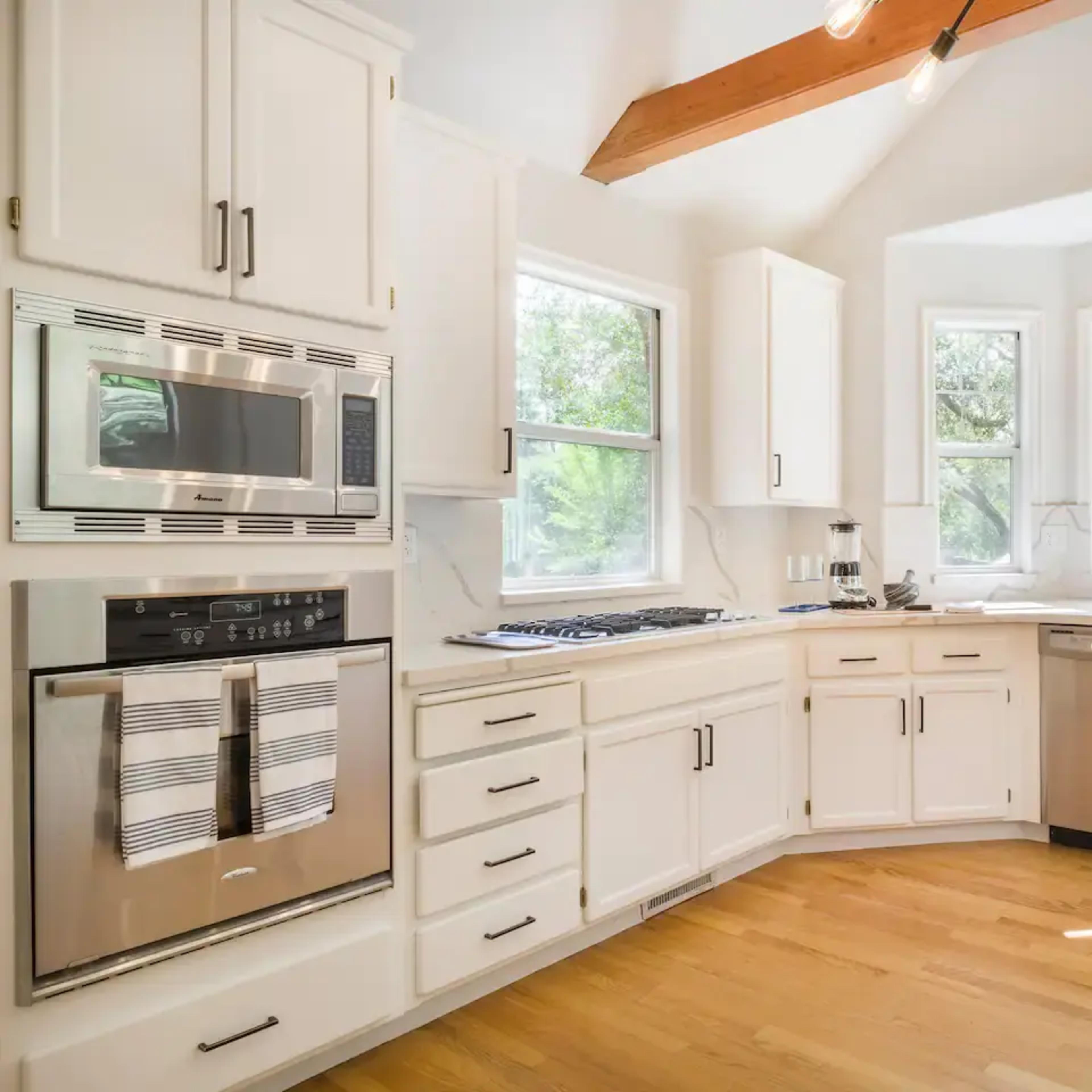 A bright kitchen features white cabinetry, stainless steel appliances, and large windows overlooking a green landscape.