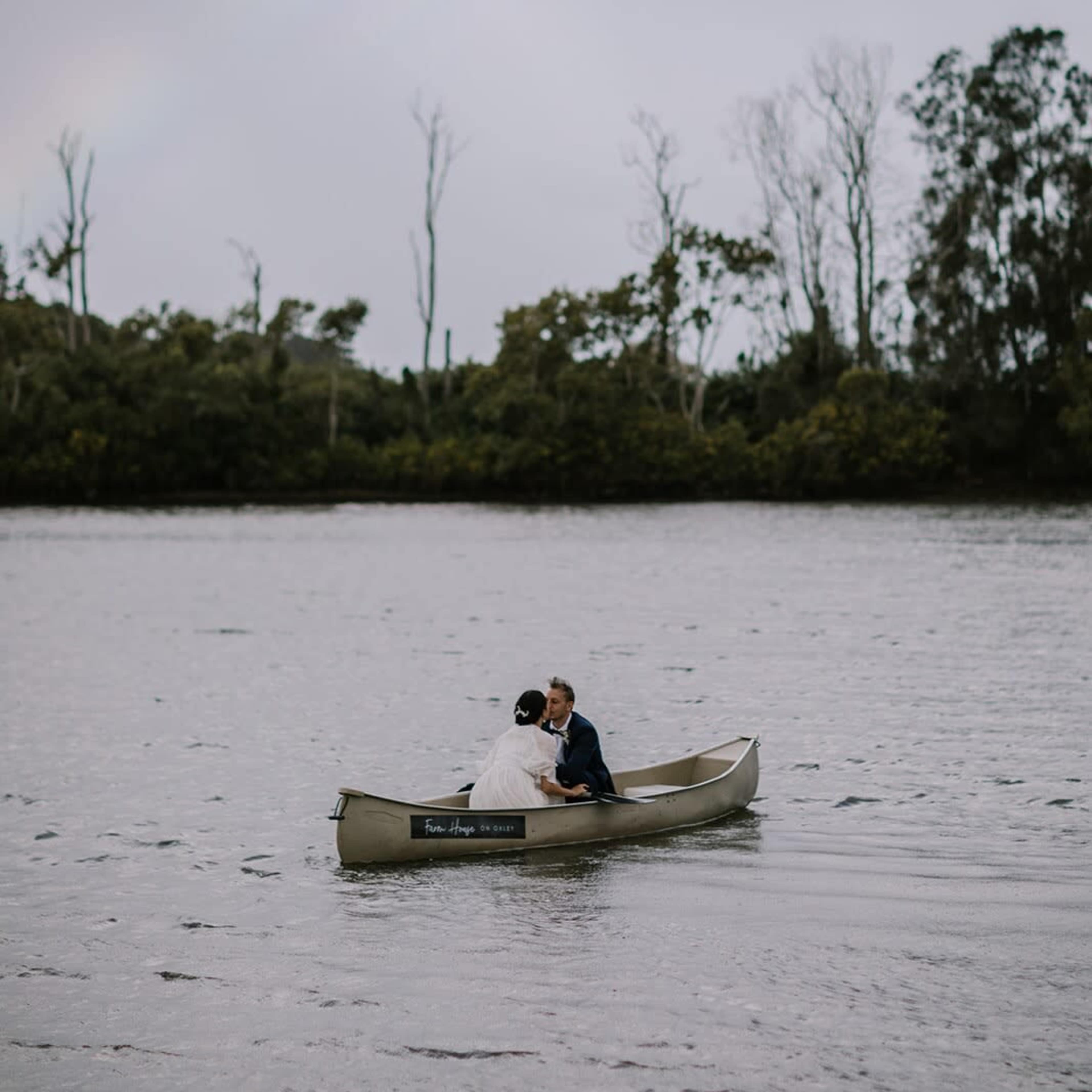 A couple in wedding attire is kissing in a canoe on a calm body of water under a rainbow.