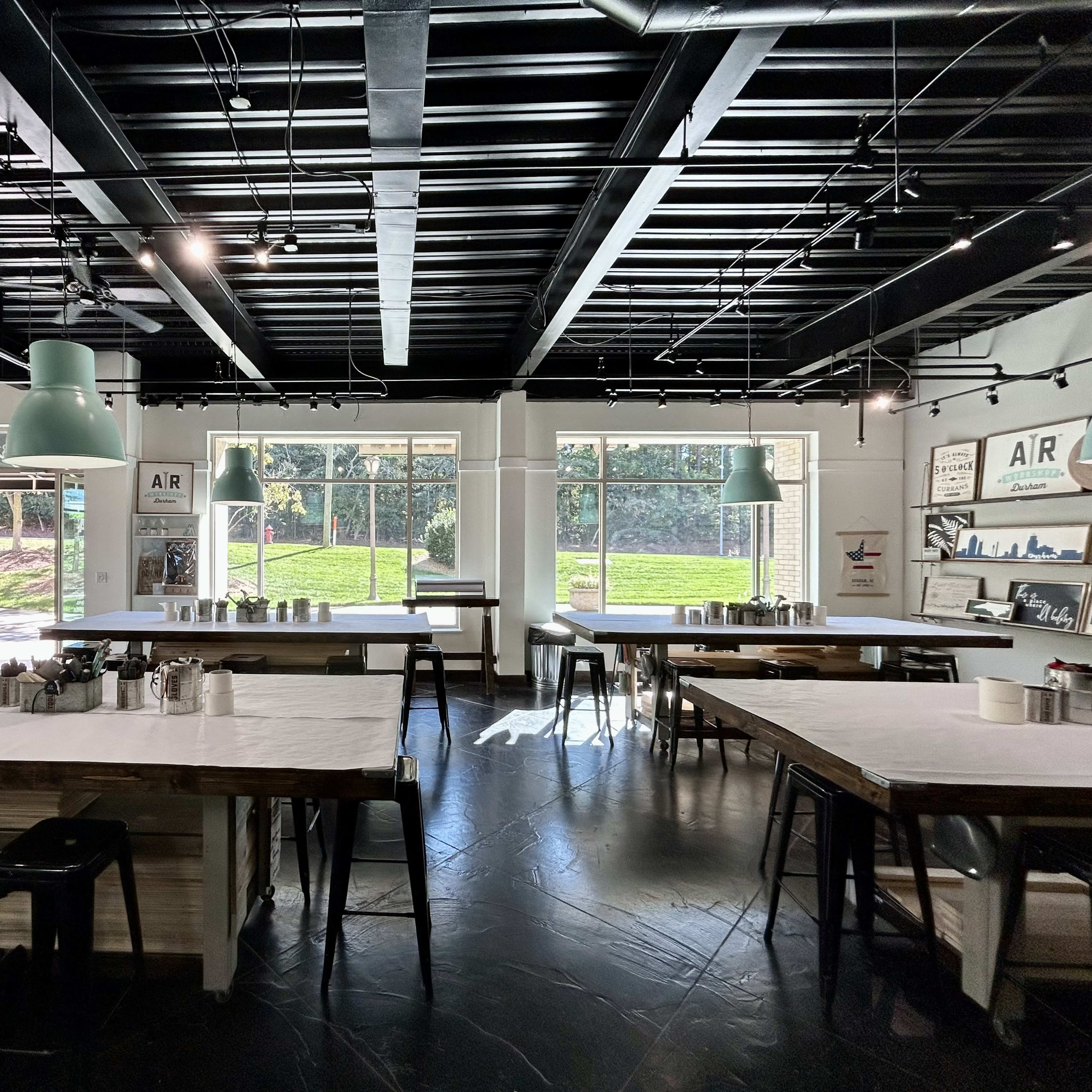 A spacious, modern café with large windows, wooden tables, and metal stools, illuminated by pendant lights.