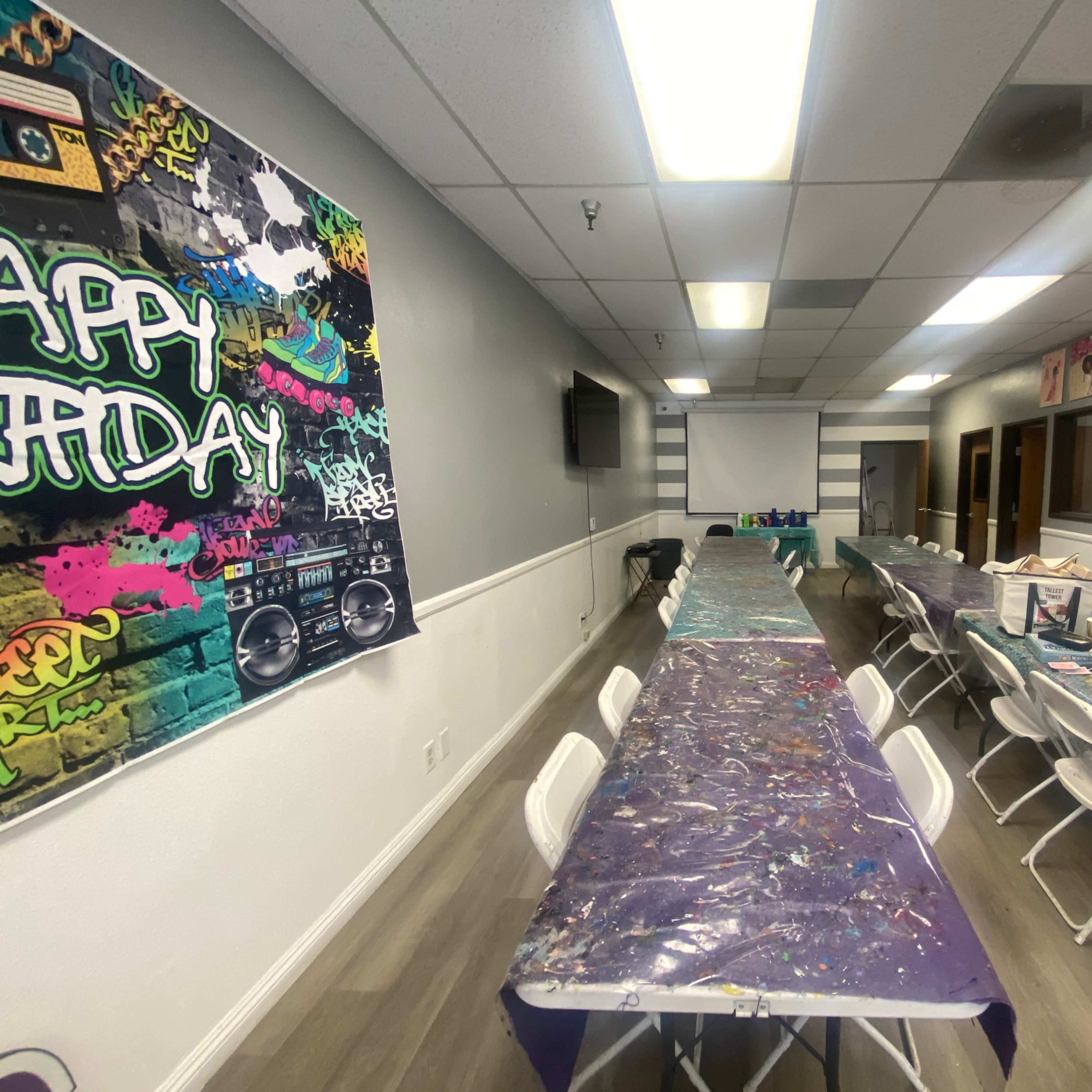 A neatly arranged event space with several long tables covered in colorful tablecloths, flanked by chairs and decorated walls featuring artwork and a "Happy Birthday" banner.