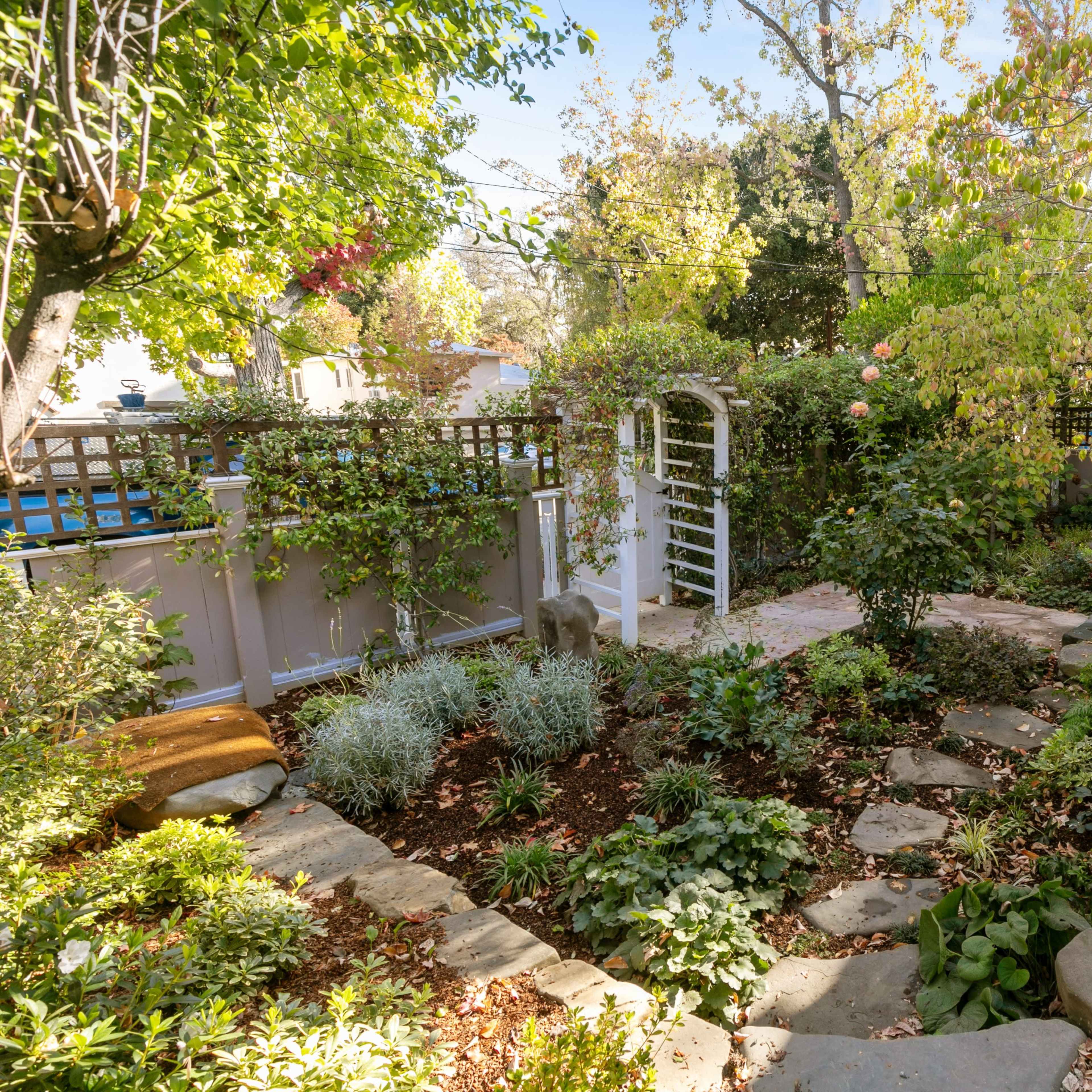 A landscaped backyard featuring a stone pathway, lush greenery, and a white archway surrounded by various plants.