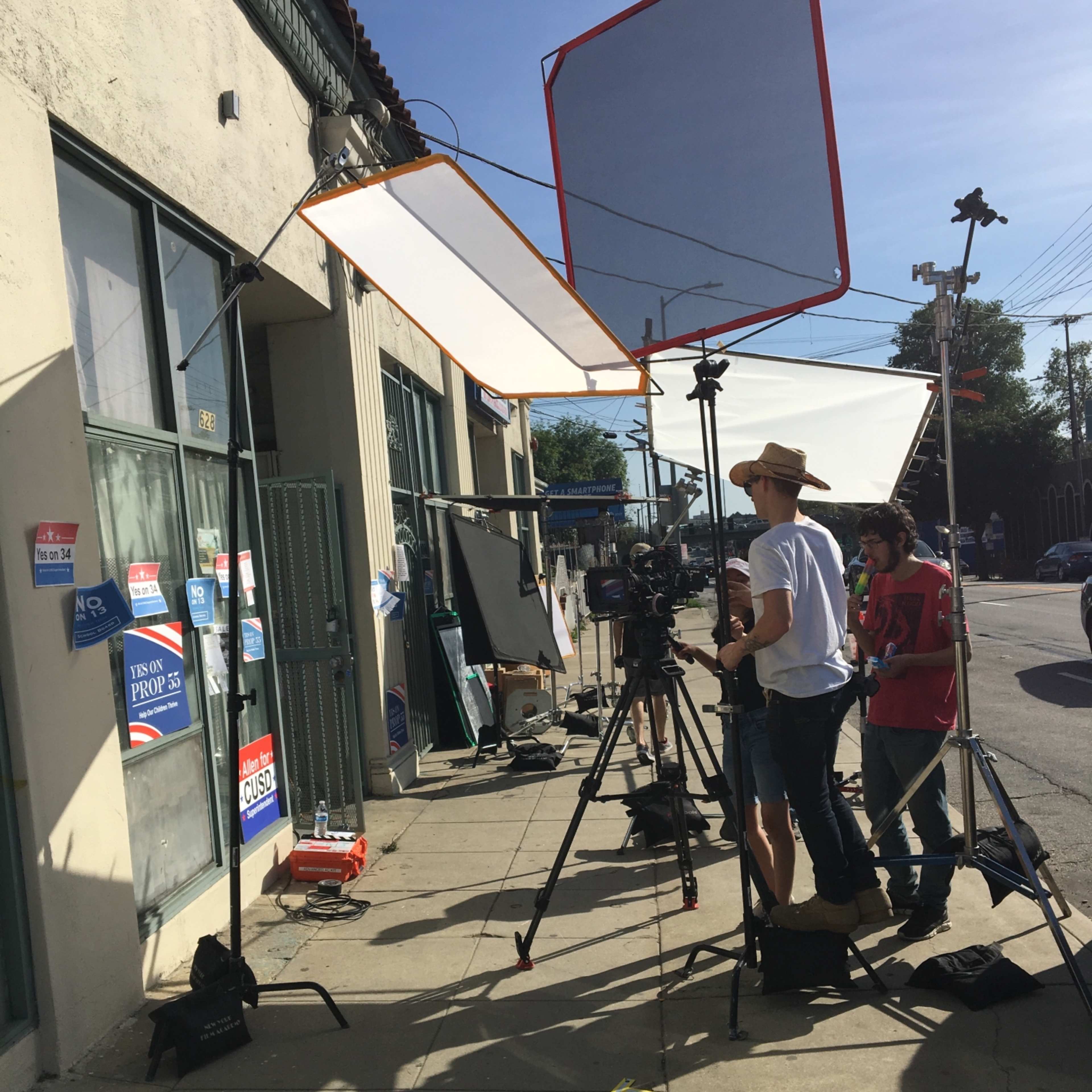 A film crew sets up equipment outside a building, with lights and cameras directed at a wall displaying various signs.