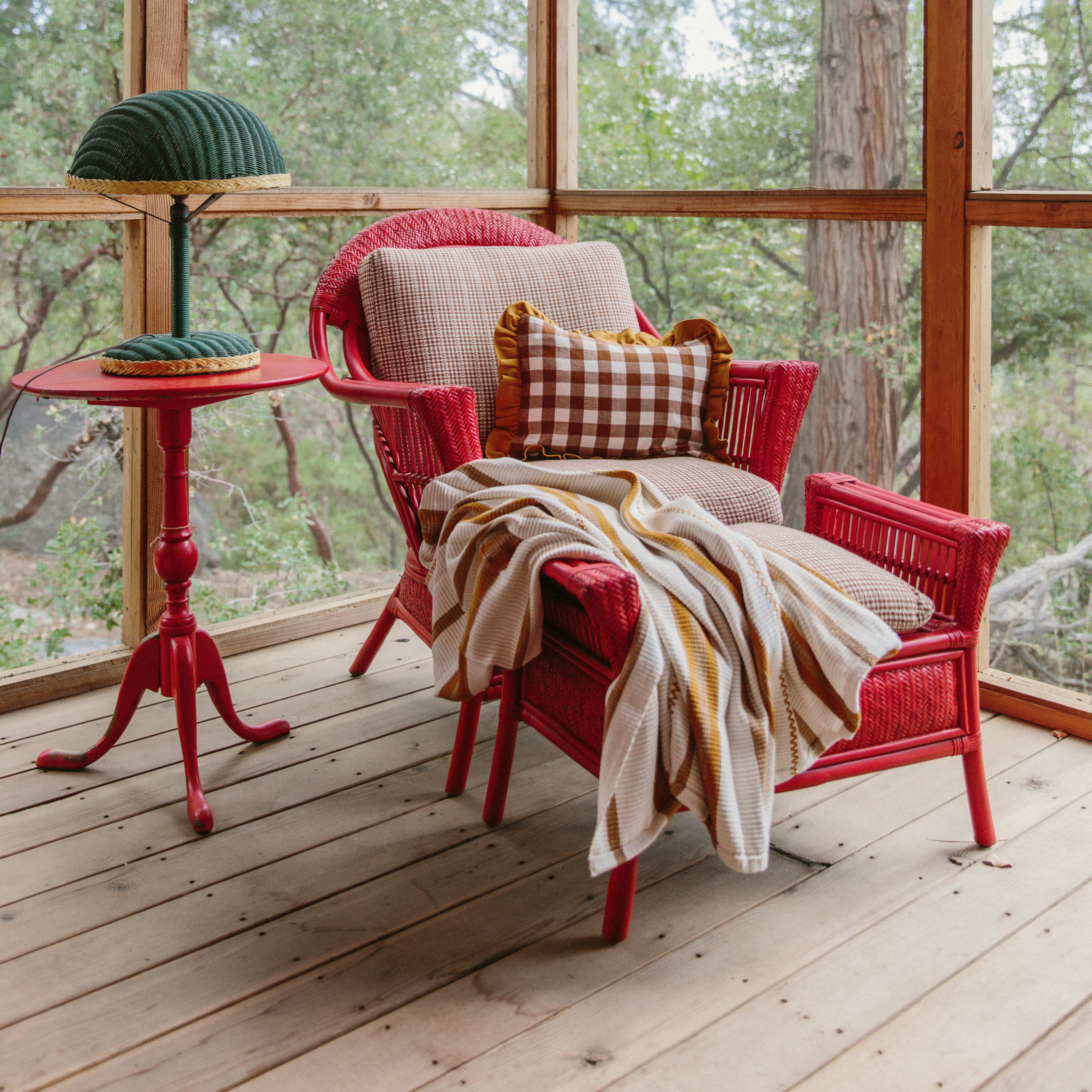 A red wicker chair with a patterned throw blanket and a small side table with a lamp is situated in a screened-in porch surrounded by trees.