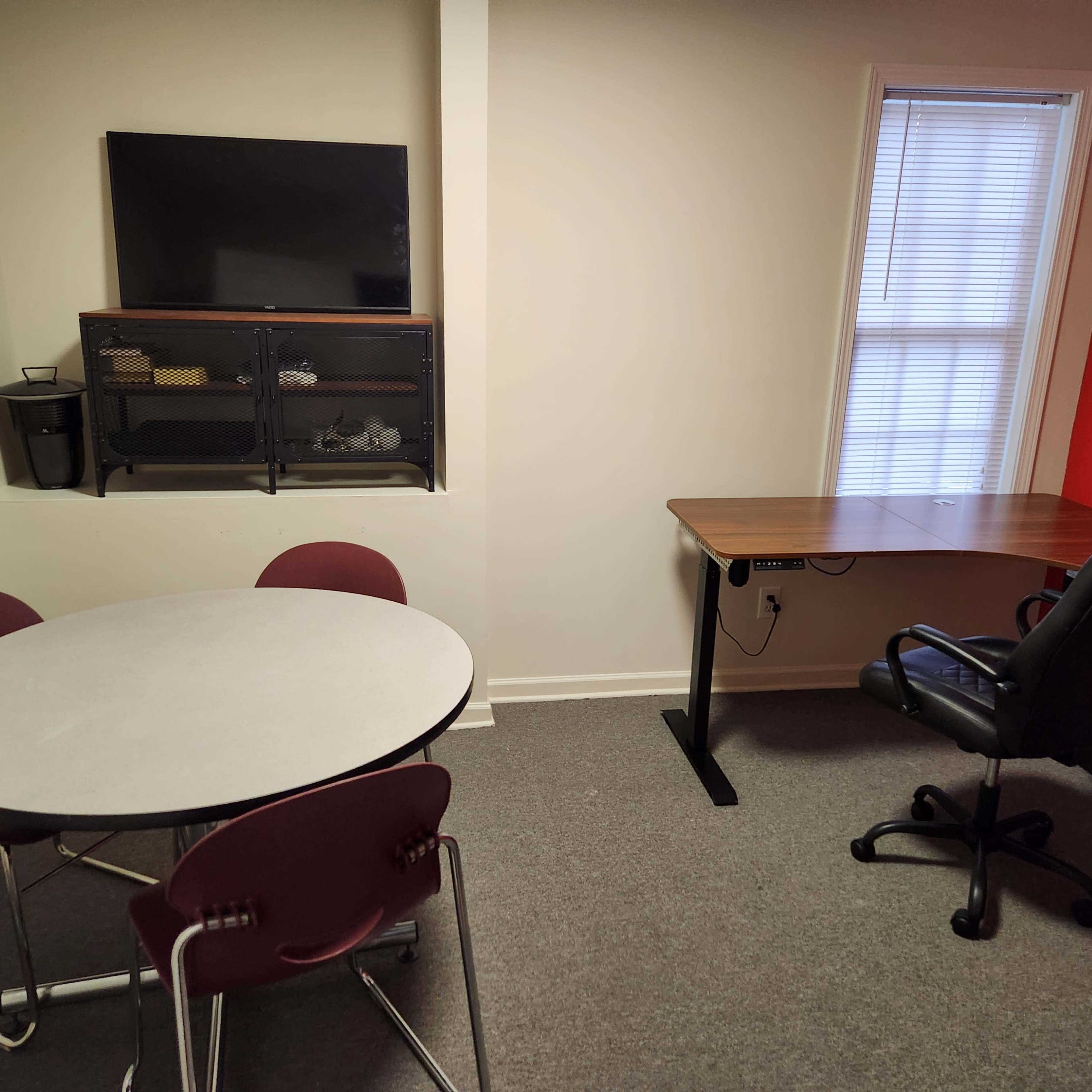 A small office space featuring a round table with four chairs, a desk with an ergonomic chair, a television mounted on the wall, and a window providing natural light.
