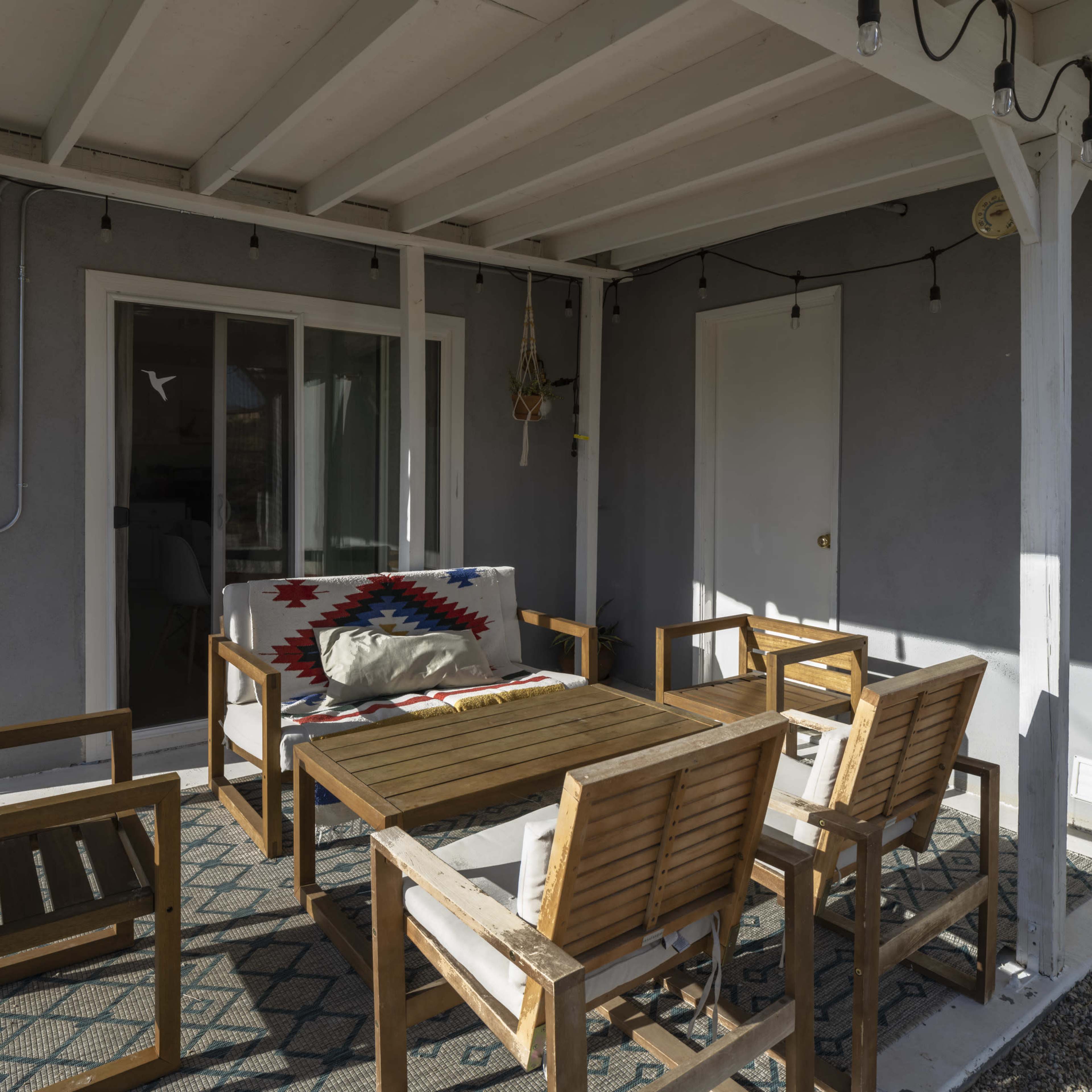 A patio area with a wooden table surrounded by four chairs, a couch with a patterned blanket, and a sliding glass door under a covered roof with string lights.