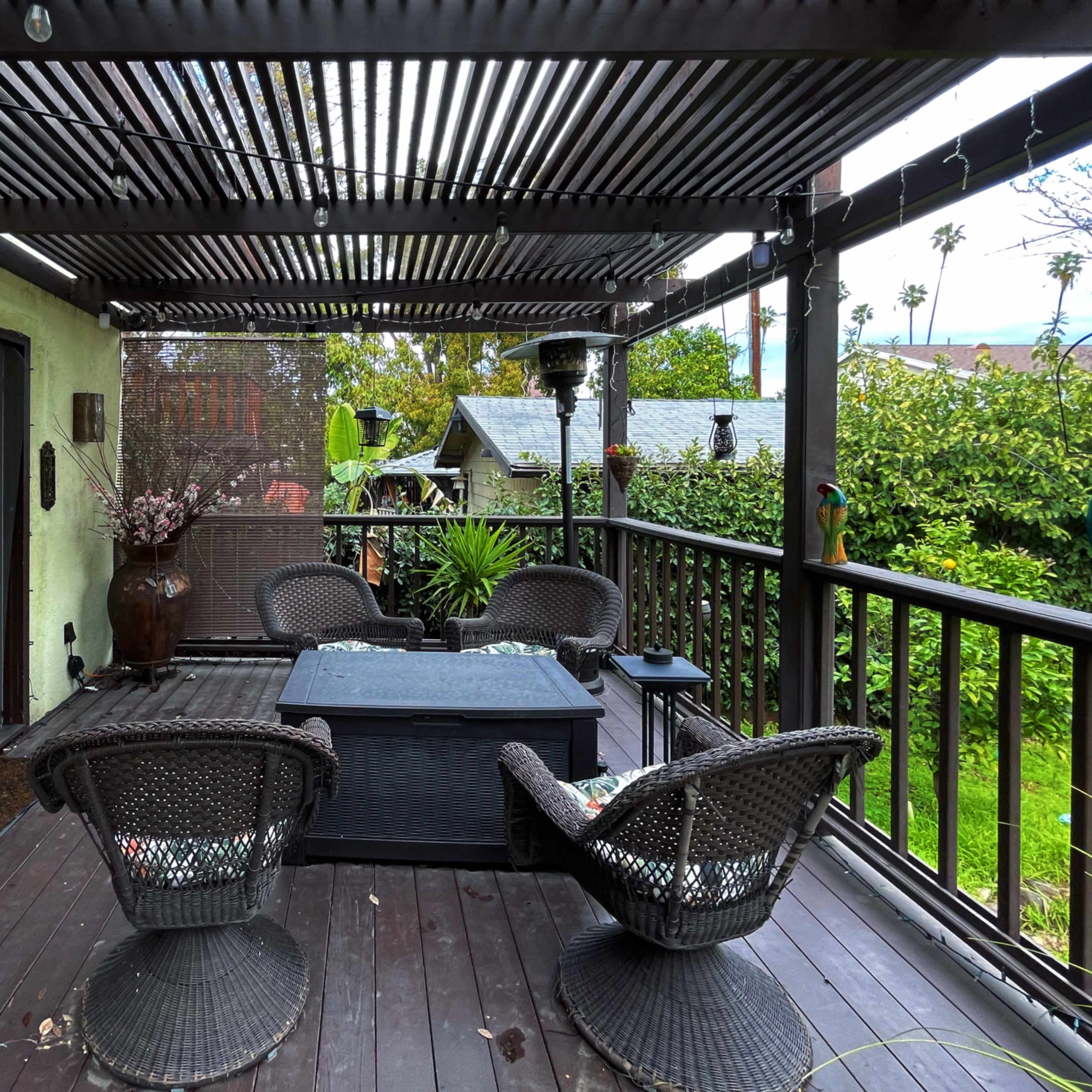 A covered patio features wicker seating around a black coffee table, surrounded by greenery and adjoining structures.