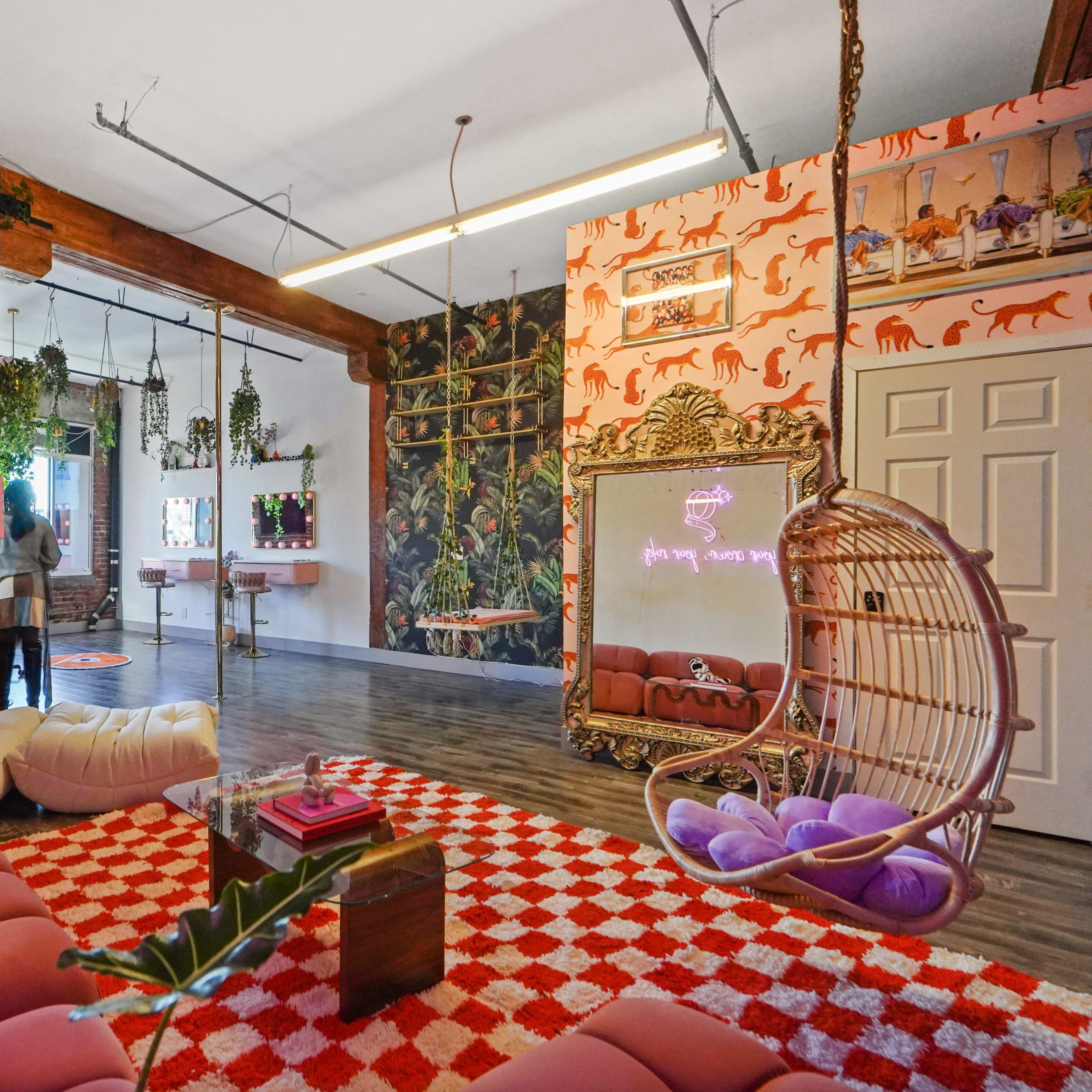A colorful room featuring a pink sofa, a patterned red and white rug, a swinging chair, and various plants along the walls.