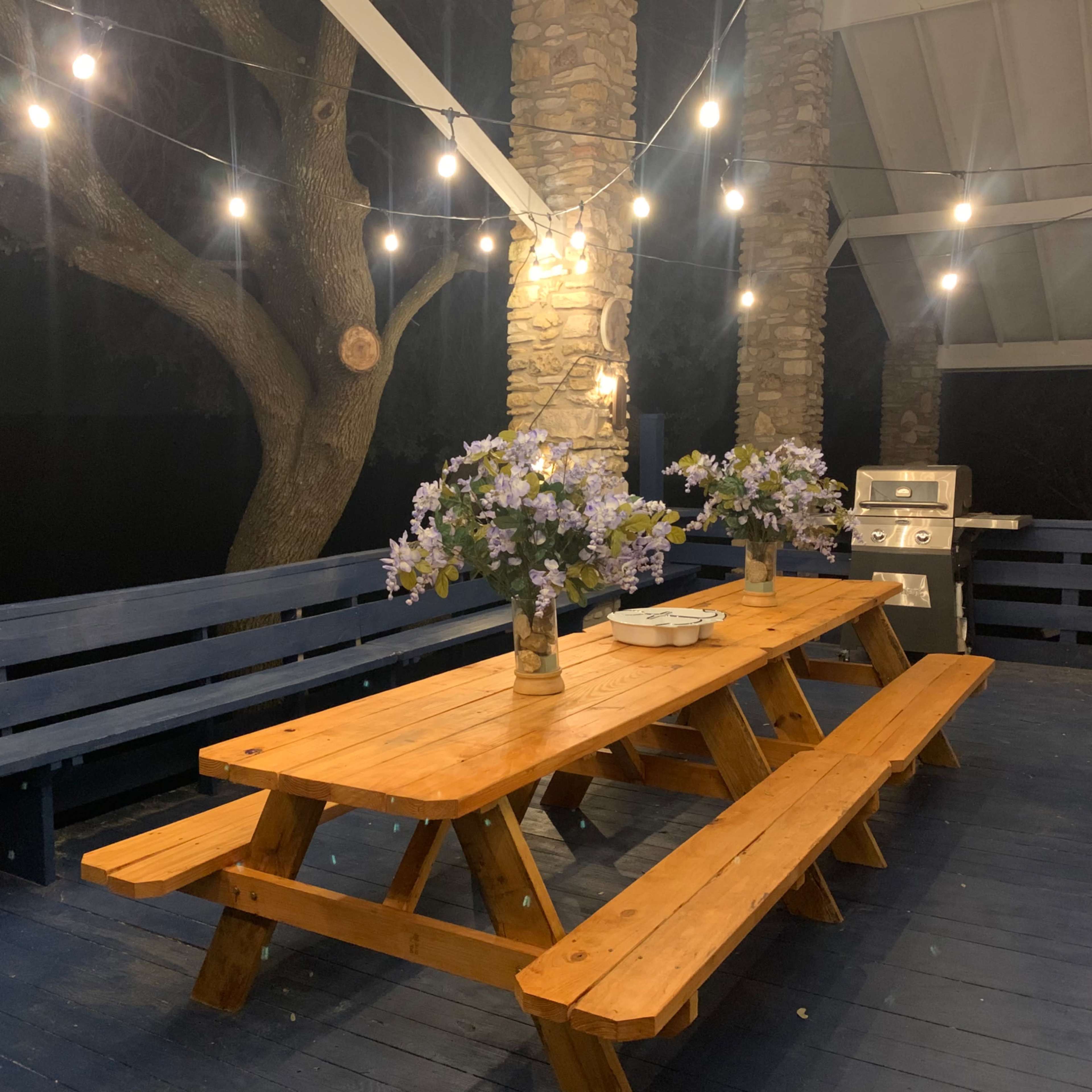 A wooden picnic table with benches is set on a deck, surrounded by stone pillars and string lights, with two vases of flowers on the table.