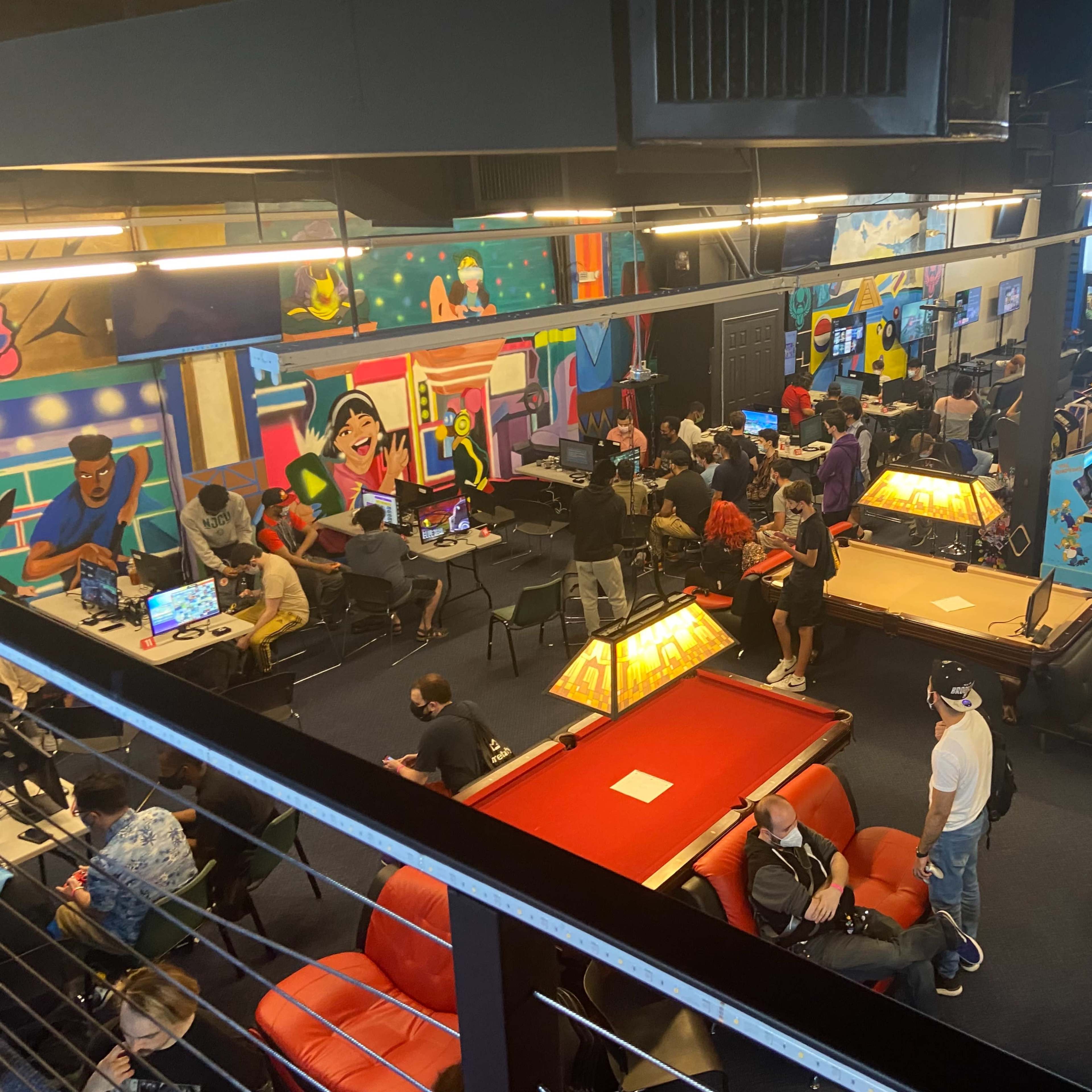 A busy gaming arcade filled with people playing various video games, with colorful murals on the walls and gaming stations set up throughout the space.