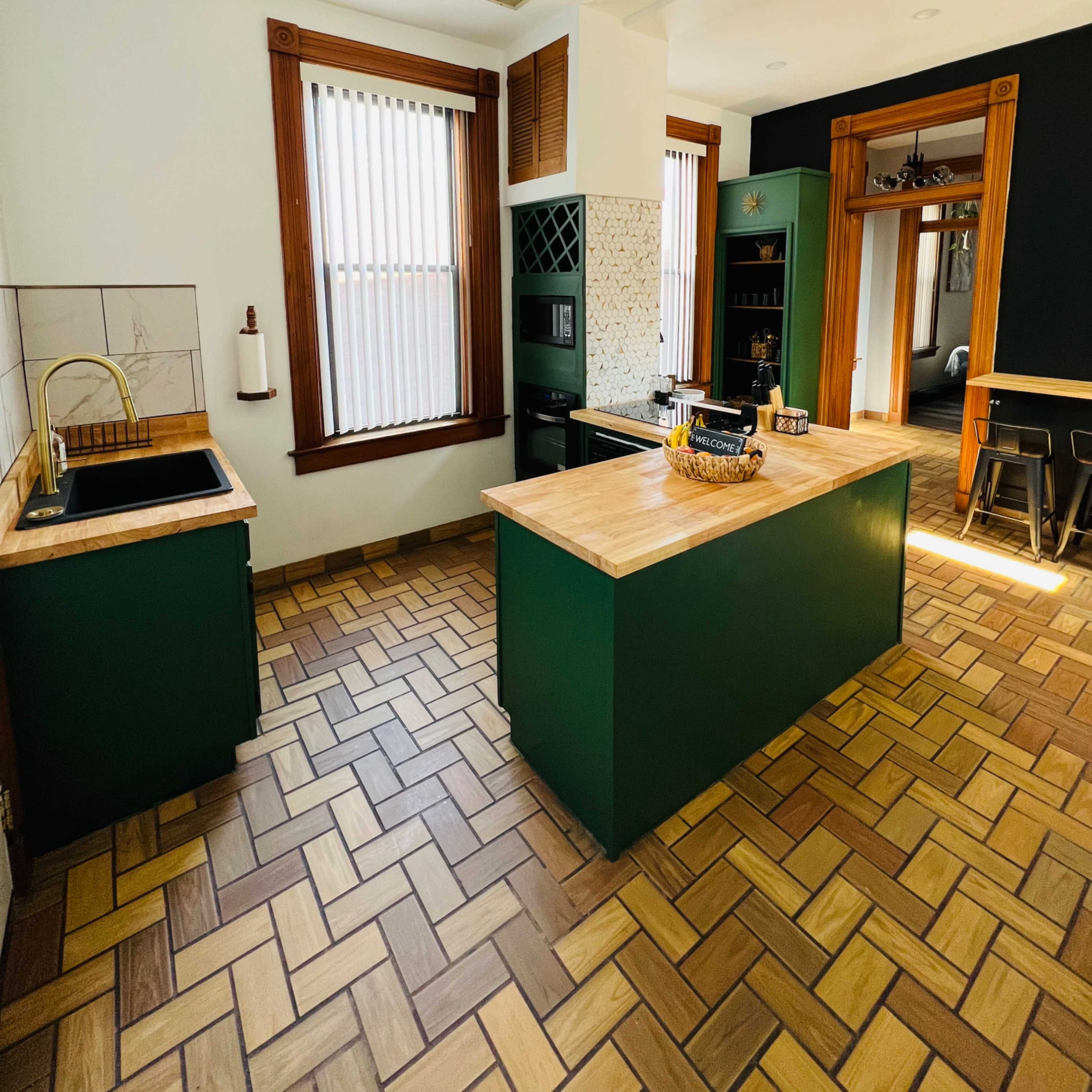 A well-lit kitchen featuring green cabinetry, a wooden island, and brown tiled flooring with a geometric pattern.