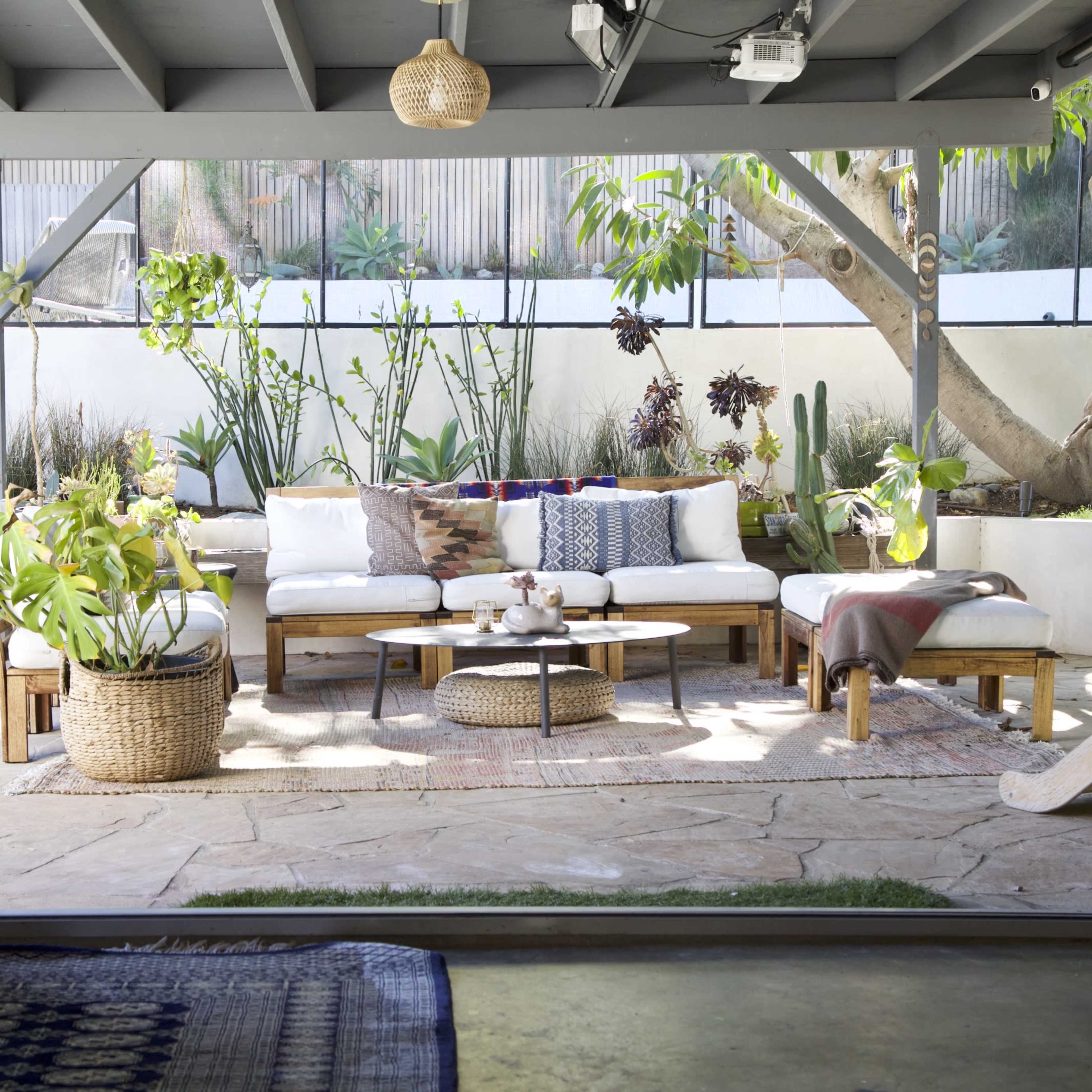 A cozy outdoor lounge area features a sectional sofa, woven coffee table, and various plants, framed by large glass doors.