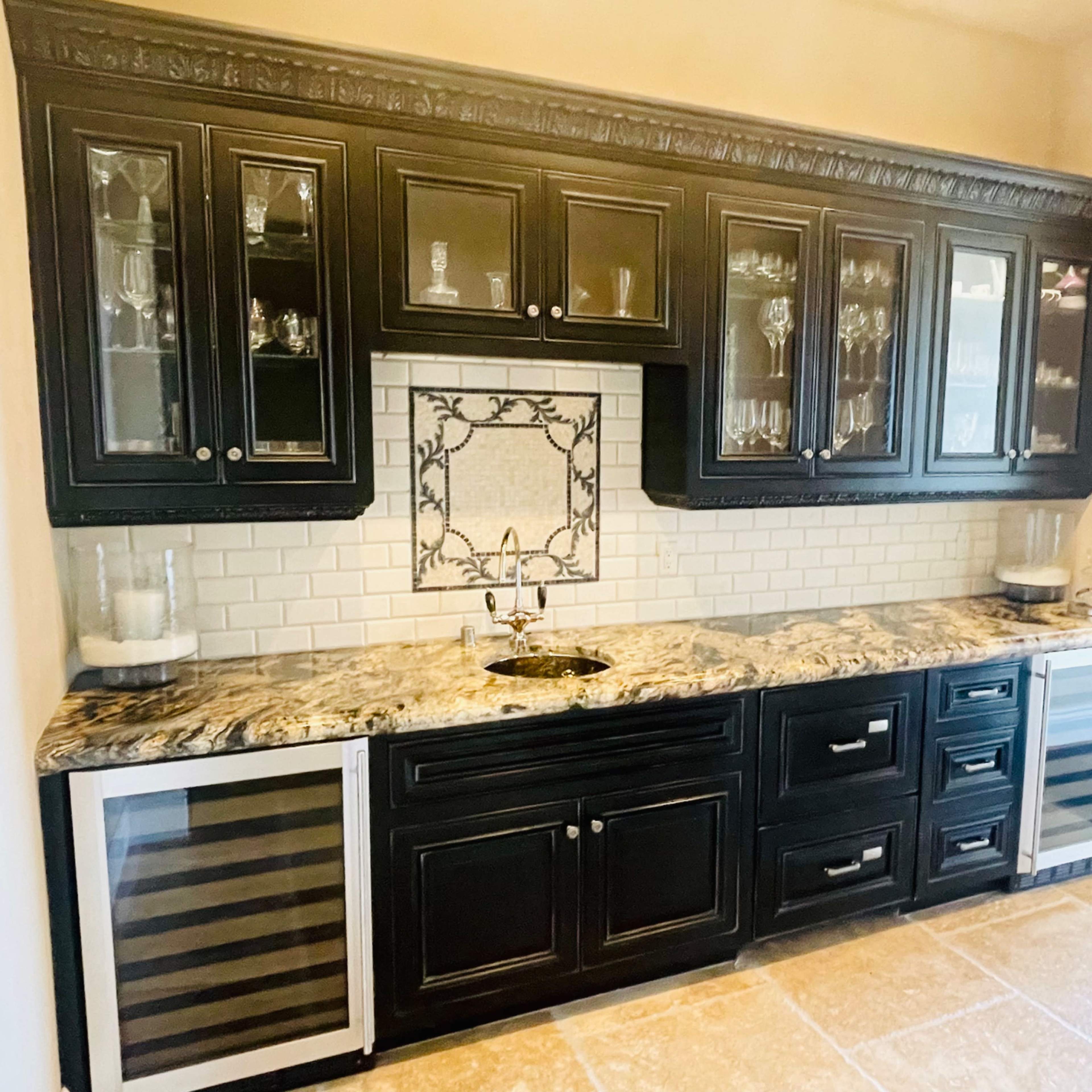 A kitchen bar area with black cabinets, a granite countertop, a backsplash featuring a decorative tile, and two wine coolers.